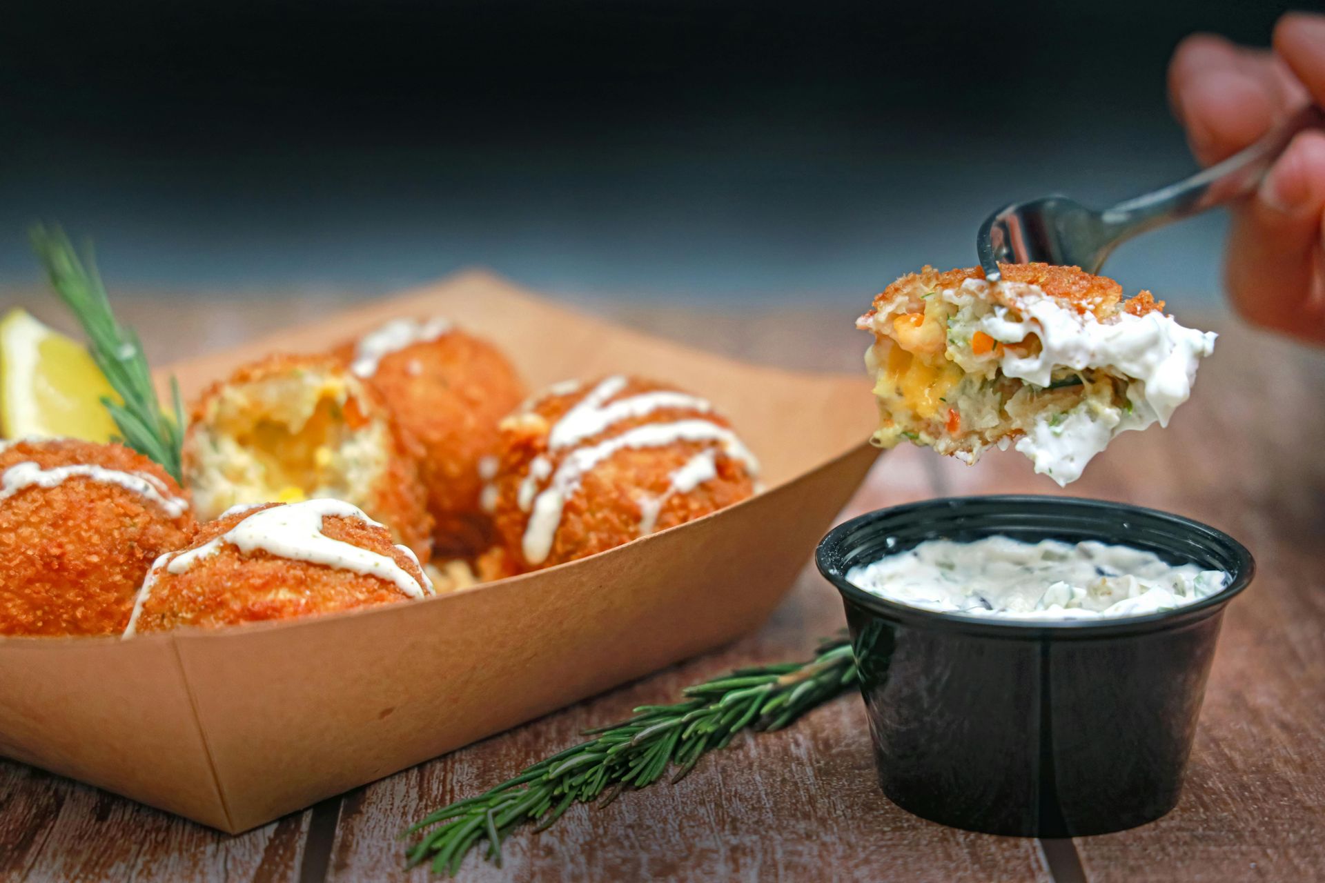 Golden fried food balls with white drizzle, served in a cardboard tray. Dipped in white sauce by a fork.