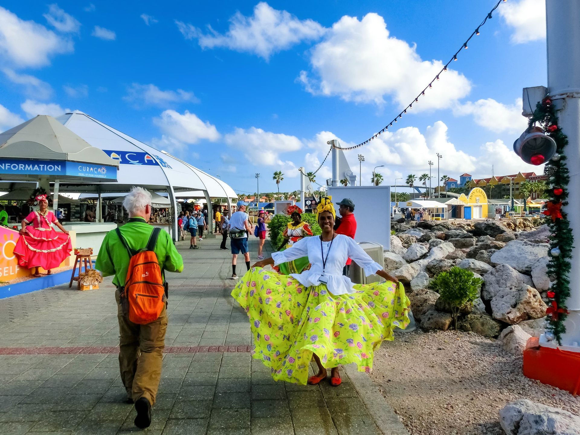 Woman in yellow dress dances on pier, blue sky, people, shops in background.