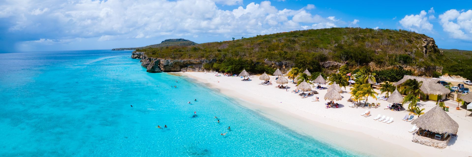 Aerial view of a white sand beach with turquoise water and thatched-roof structures surrounded by lush greenery under a partly cloudy sky.