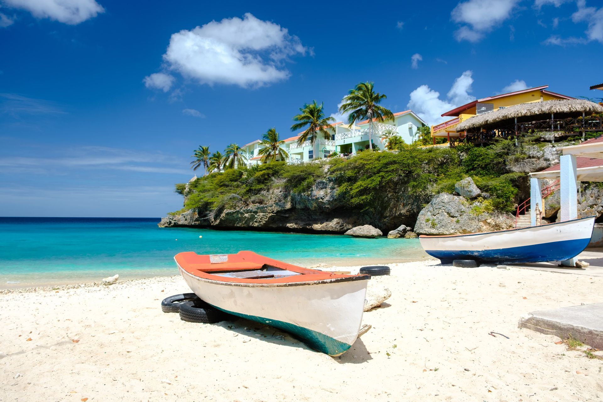 Two boats on a white sand beach, turquoise water, and buildings on a cliff under a blue sky.