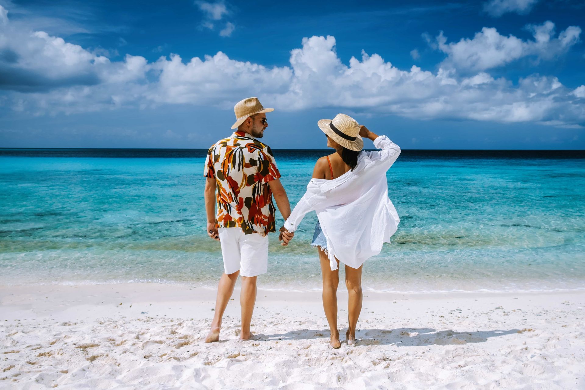 Couple holding hands, standing on a white sand beach, ocean in background. Wearing casual summer clothes.