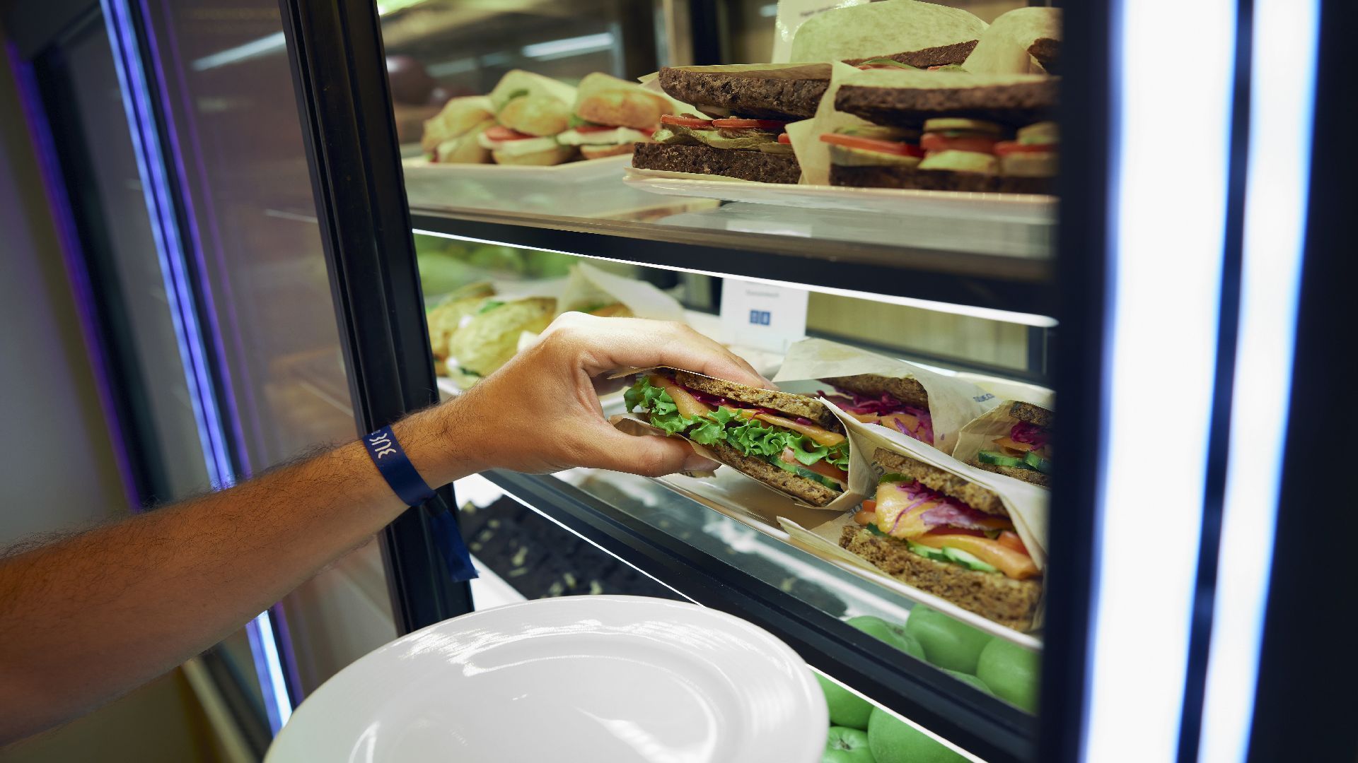 Person's hand reaching into a deli cooler for a sandwich, holding a plate.