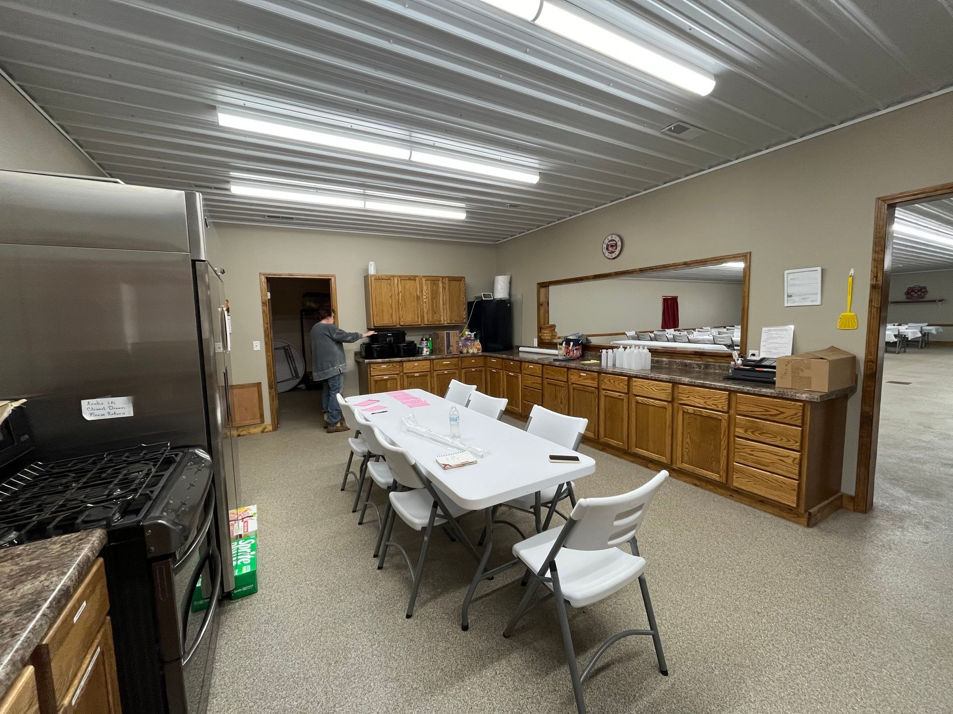 Commercial kitchen or break room with folding tables, white chairs, wooden cabinets, and fluorescent lights