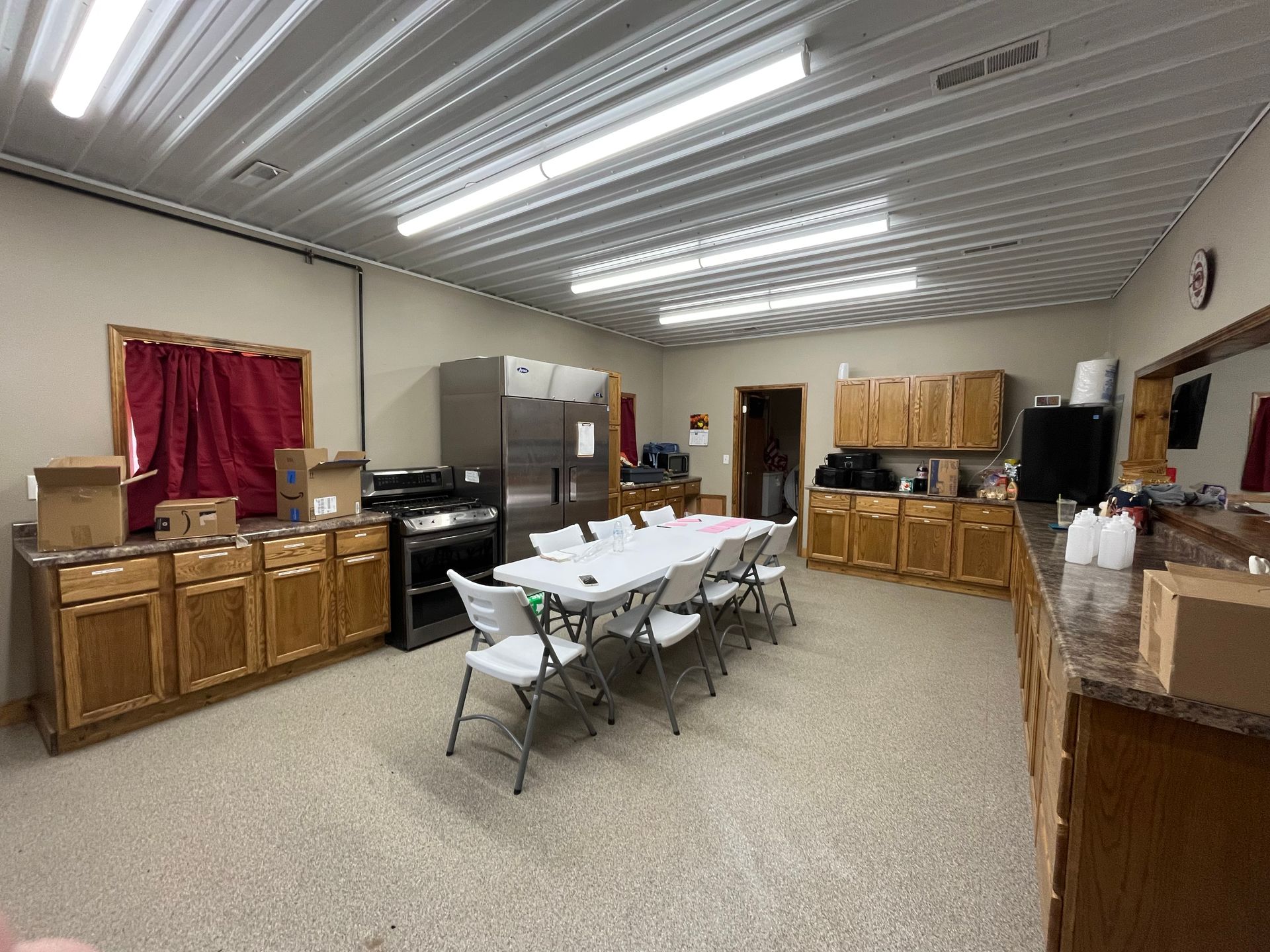 Community room with folding tables and chairs, wooden cabinets, fluorescent lights, and a red curtained window