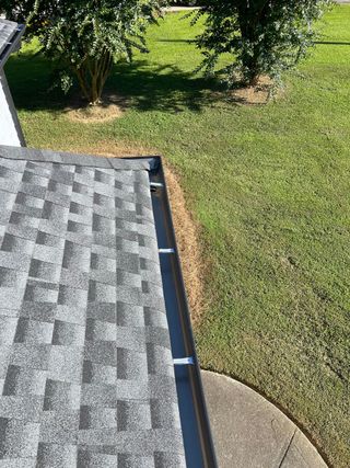 Roof edge beside a green lawn and trees, with a gutter and a round concrete pad below