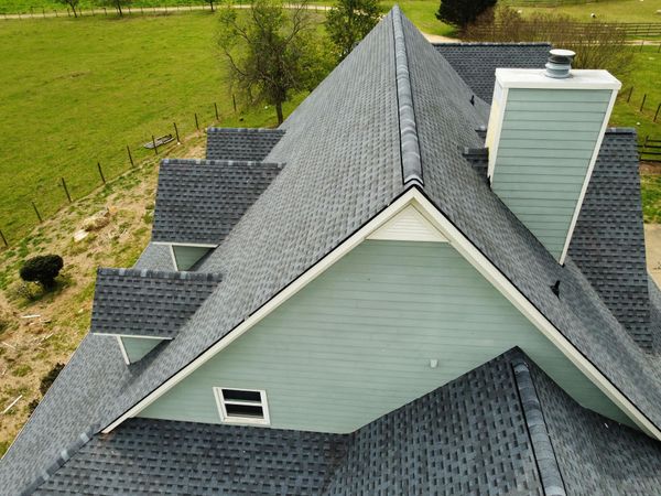 Aerial view of a gray-shingled house roof with dormers and a chimney beside a grassy field.