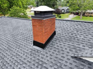 Gray shingle roof with a red brick chimney and black cap, seen against a green neighborhood backdrop