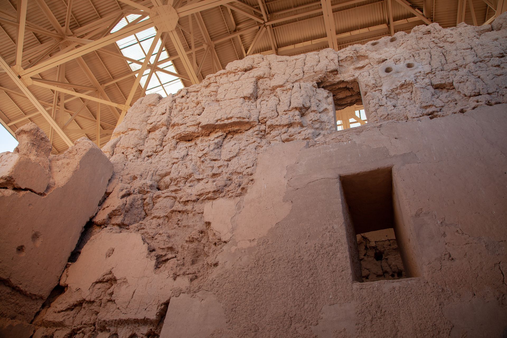 Ruins of an adobe wall under a wooden roof, with a small rectangular opening in the foreground