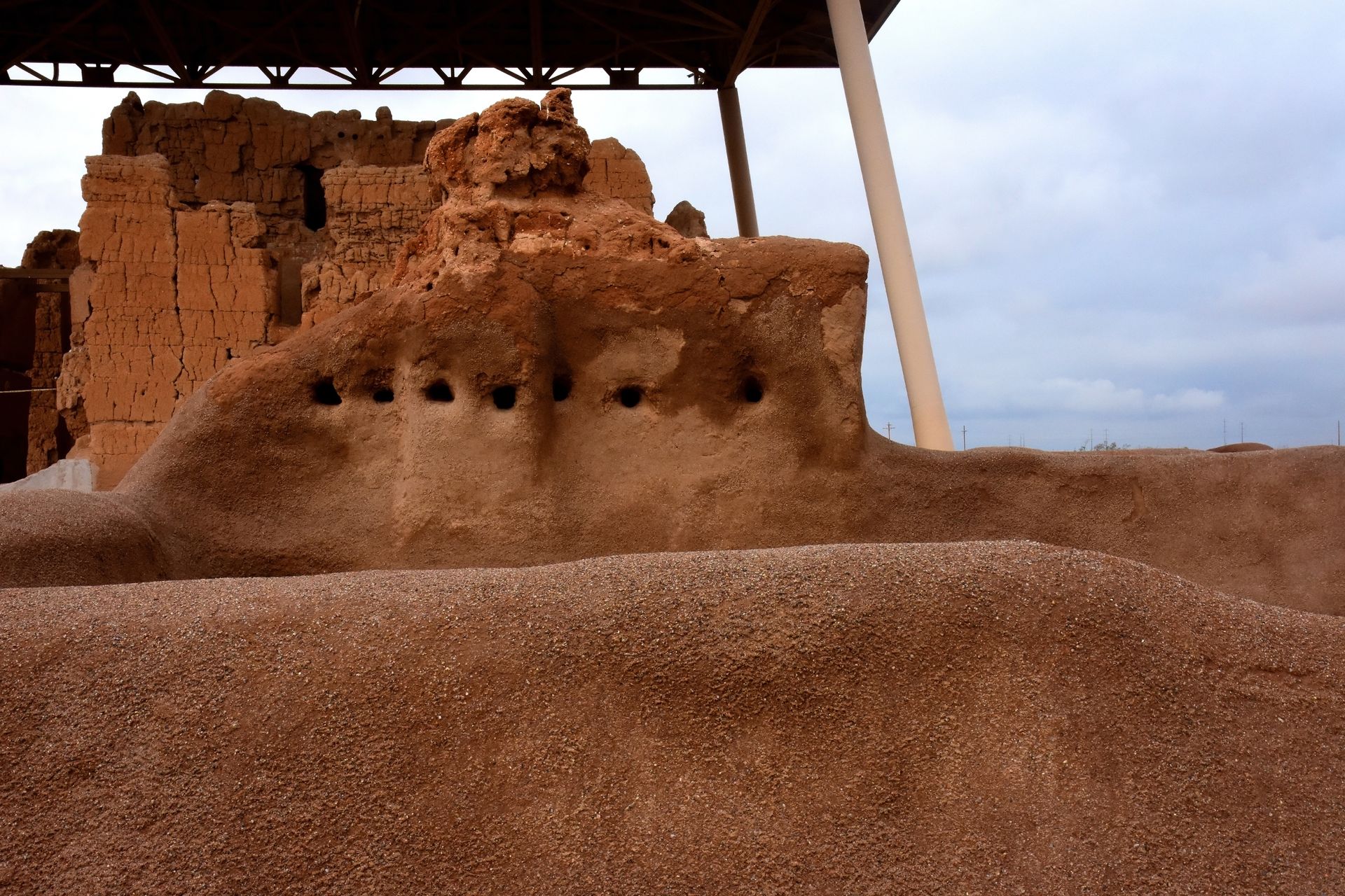Ancient adobe ruins under a shelter, with a row of small round holes in the wall and a cloudy sky behind.