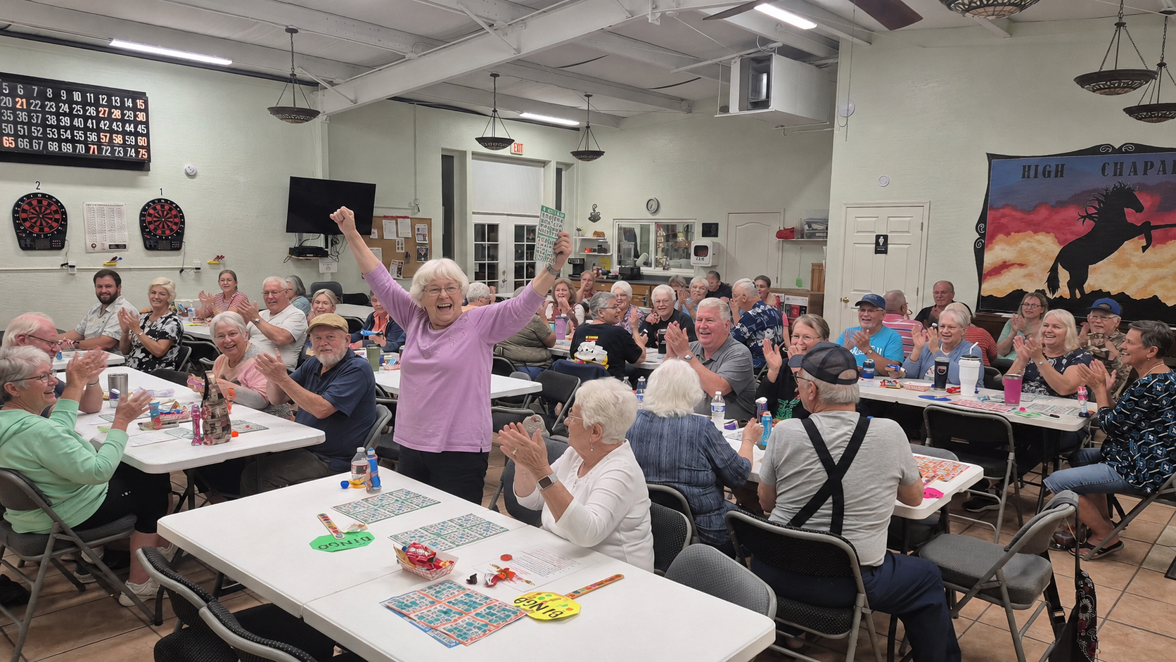 Community hall full of seated people with cards on tables; one woman in pink raises her arms near the front