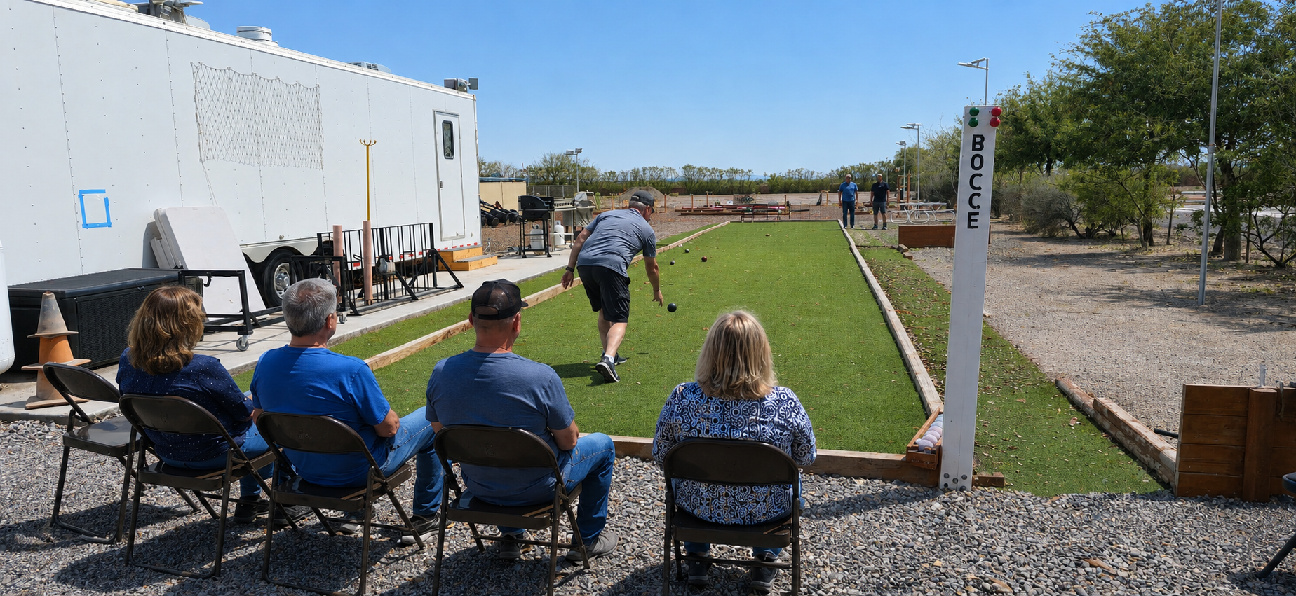 People seated on folding chairs watching a lawn game outdoors near a white trailer.