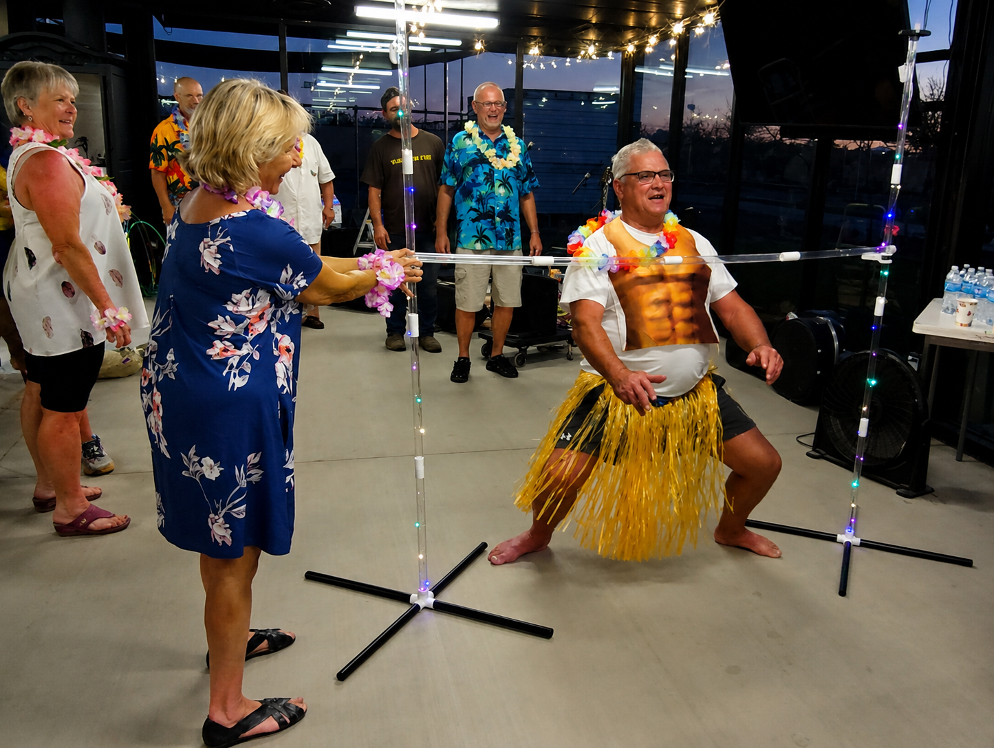 Two people pose in colorful costumes indoors, one crouching on a tripod stand while others watch nearby.