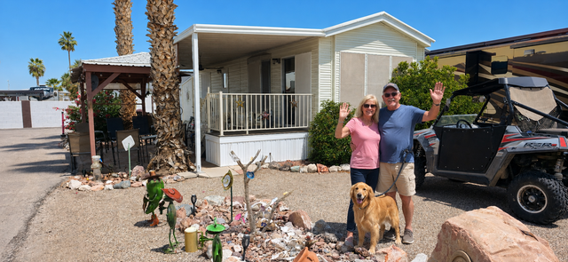 Two people and a dog posing by an RV outside a mobile home in a desert park, waving at the camera