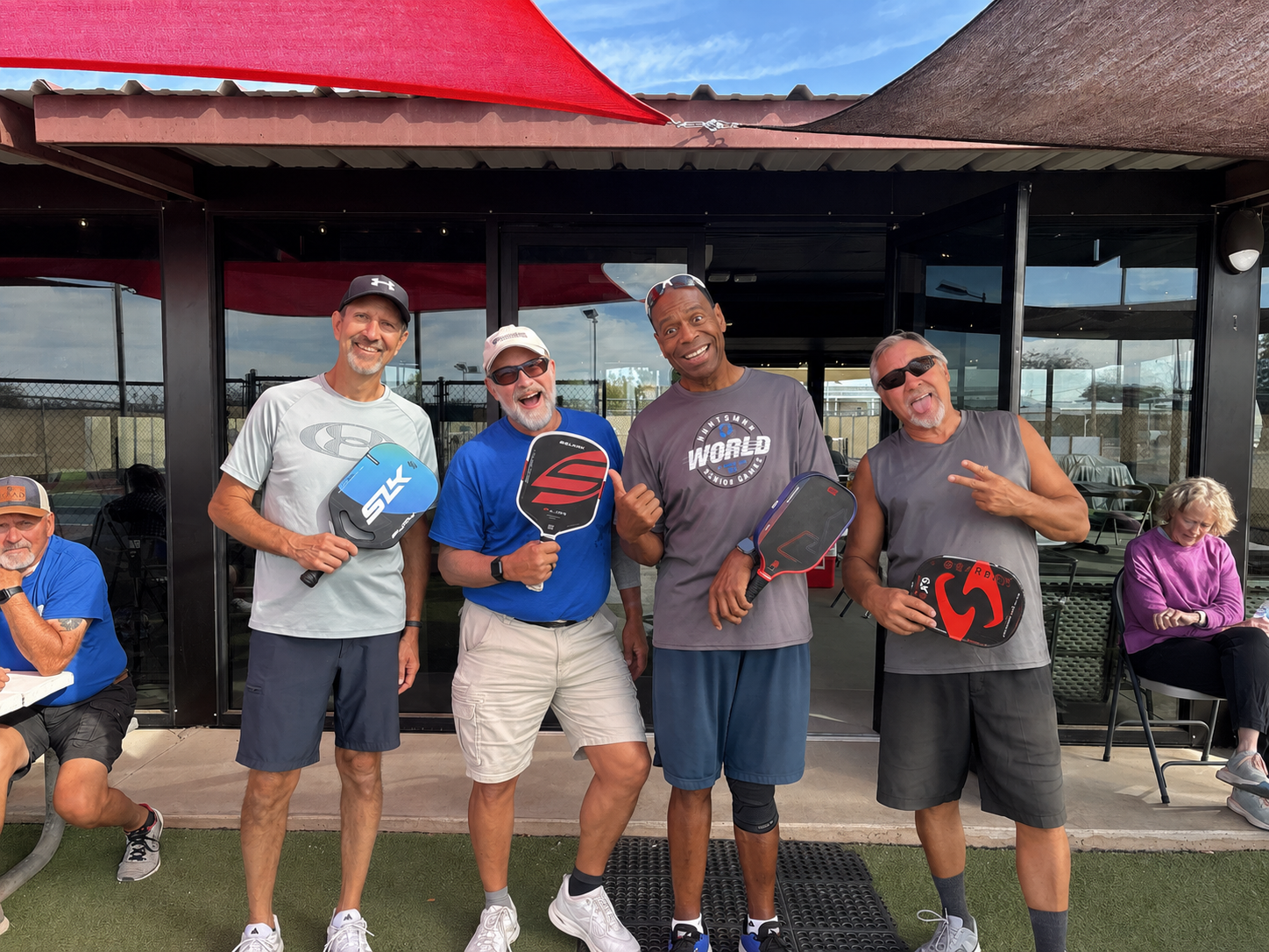 Five people posing with paddles outside a waterfront building, smiling and holding up gear