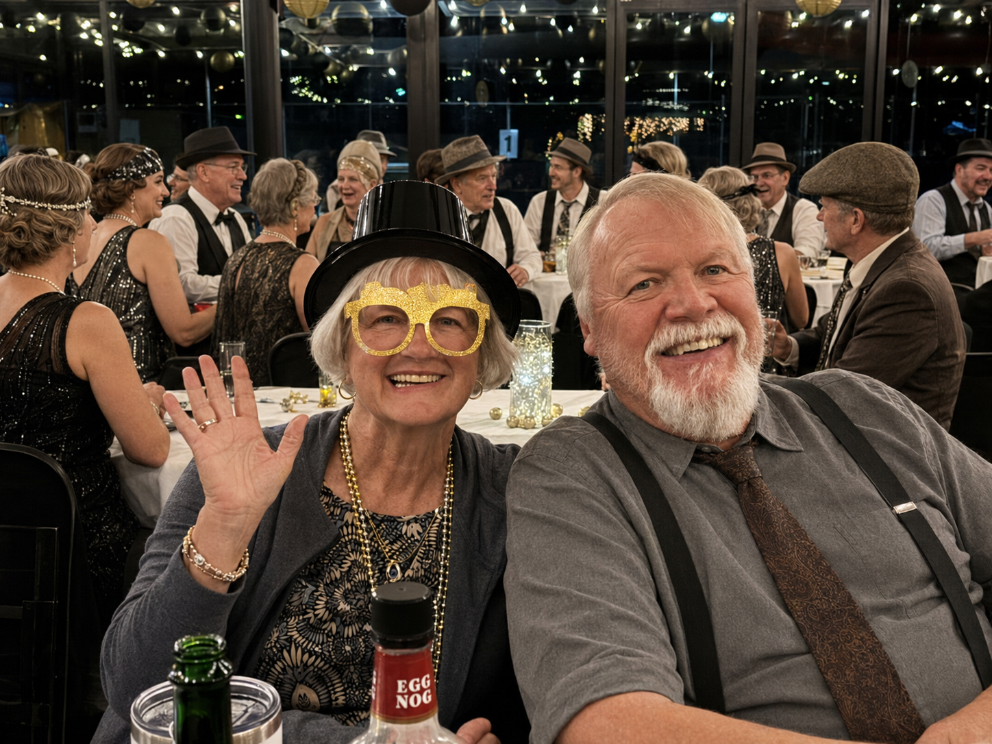 Smiling couple at a formal dinner, waving at the camera, with glasses and bottles on the table.