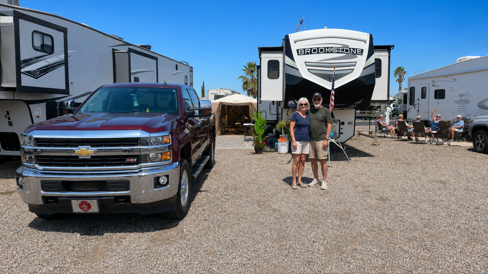 Black pickup truck and two people standing in front of a white RV in a gravel RV park under a blue sky