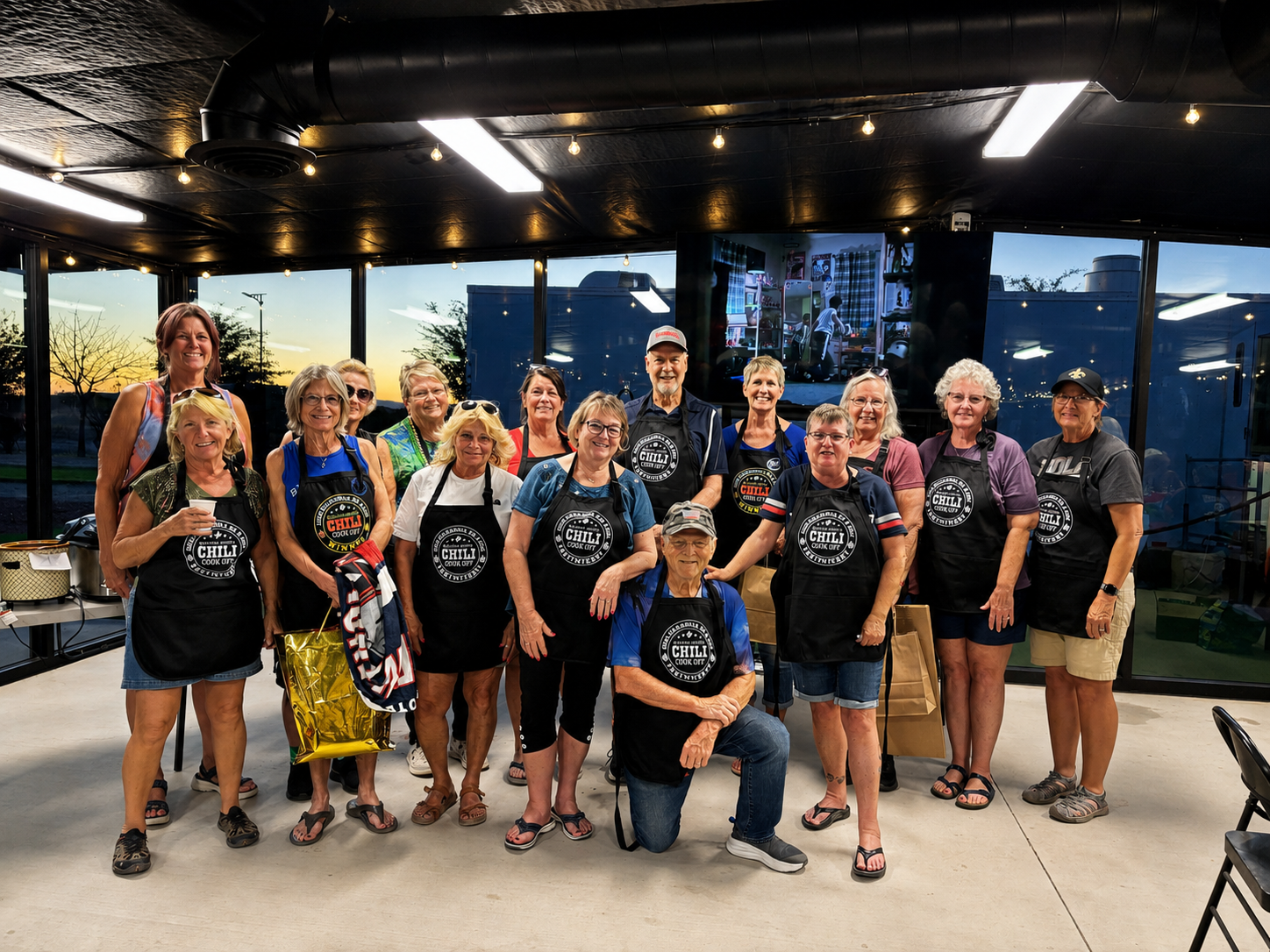 Group of people posing together indoors at a casual event, some holding drinks and wearing matching black shirts.