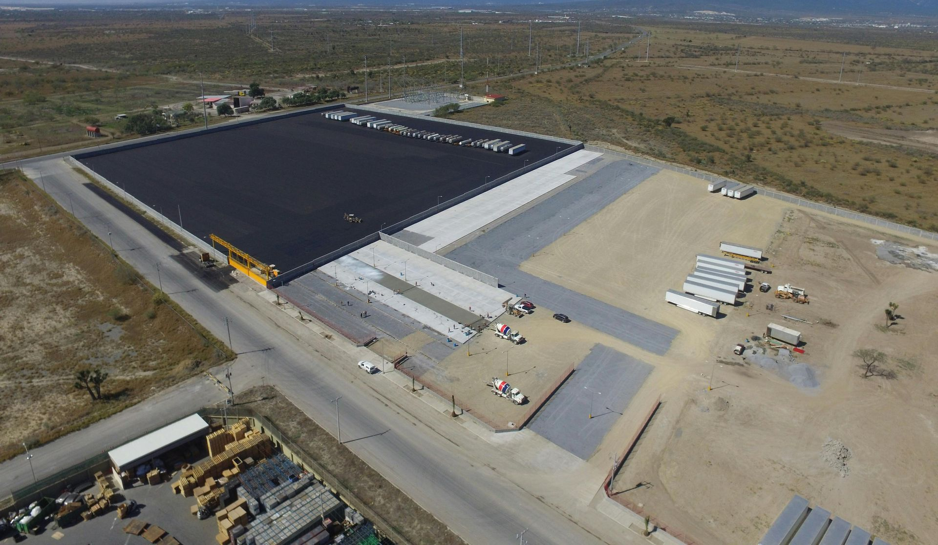 Aerial view of a large industrial site with a black-roofed warehouse, parking areas, and construction materials
