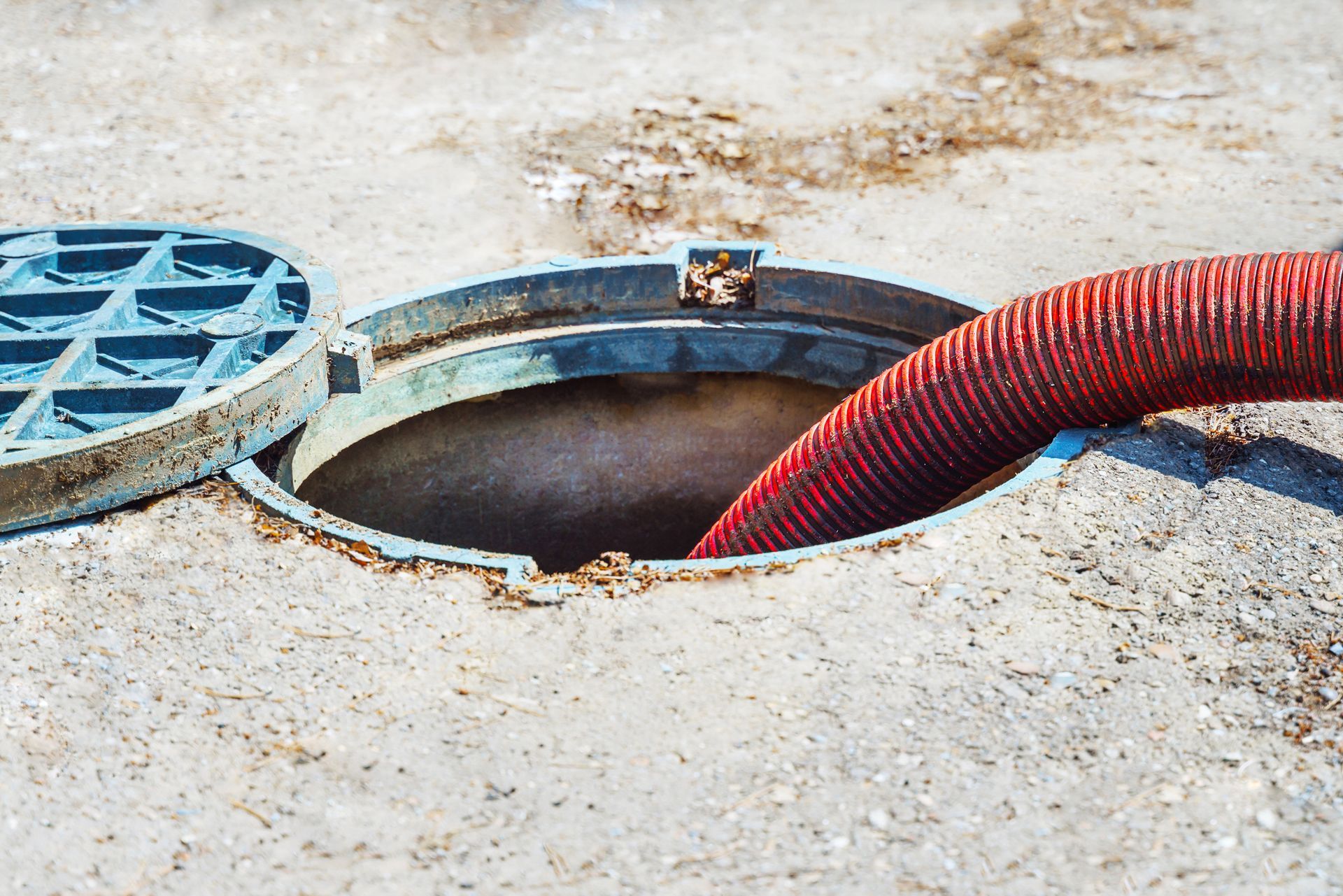 Red hose in open manhole during urban sewer maintenance on concrete surface with cover removed.