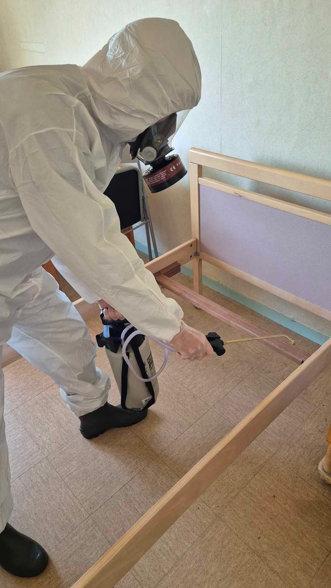 Un homme en combinaison de protection pulvérise un cadre de lit avec un pulvérisateur.