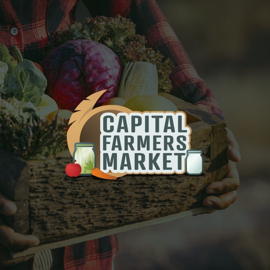 A person is holding a wooden box full of vegetables and the logo for capital farmers market