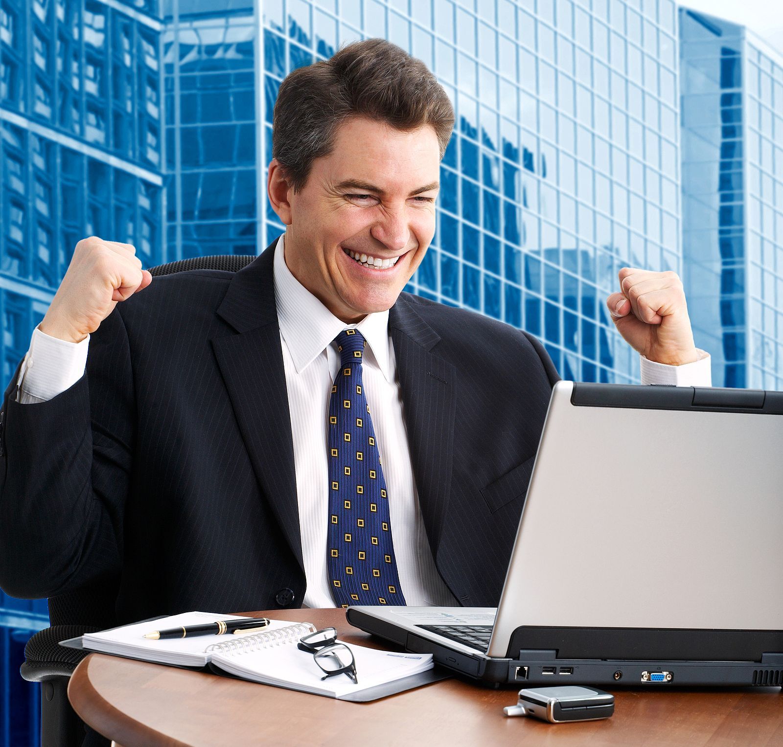 Smiling businessman in suit with raised fists at desk beside laptop in glass office