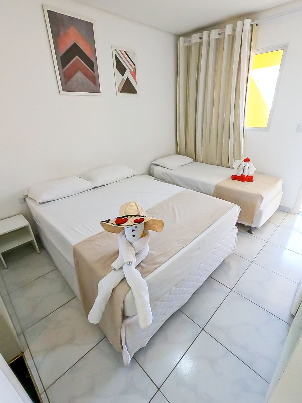 Um quarto com duas camas, uma mesa de cabeceira e uma janela.