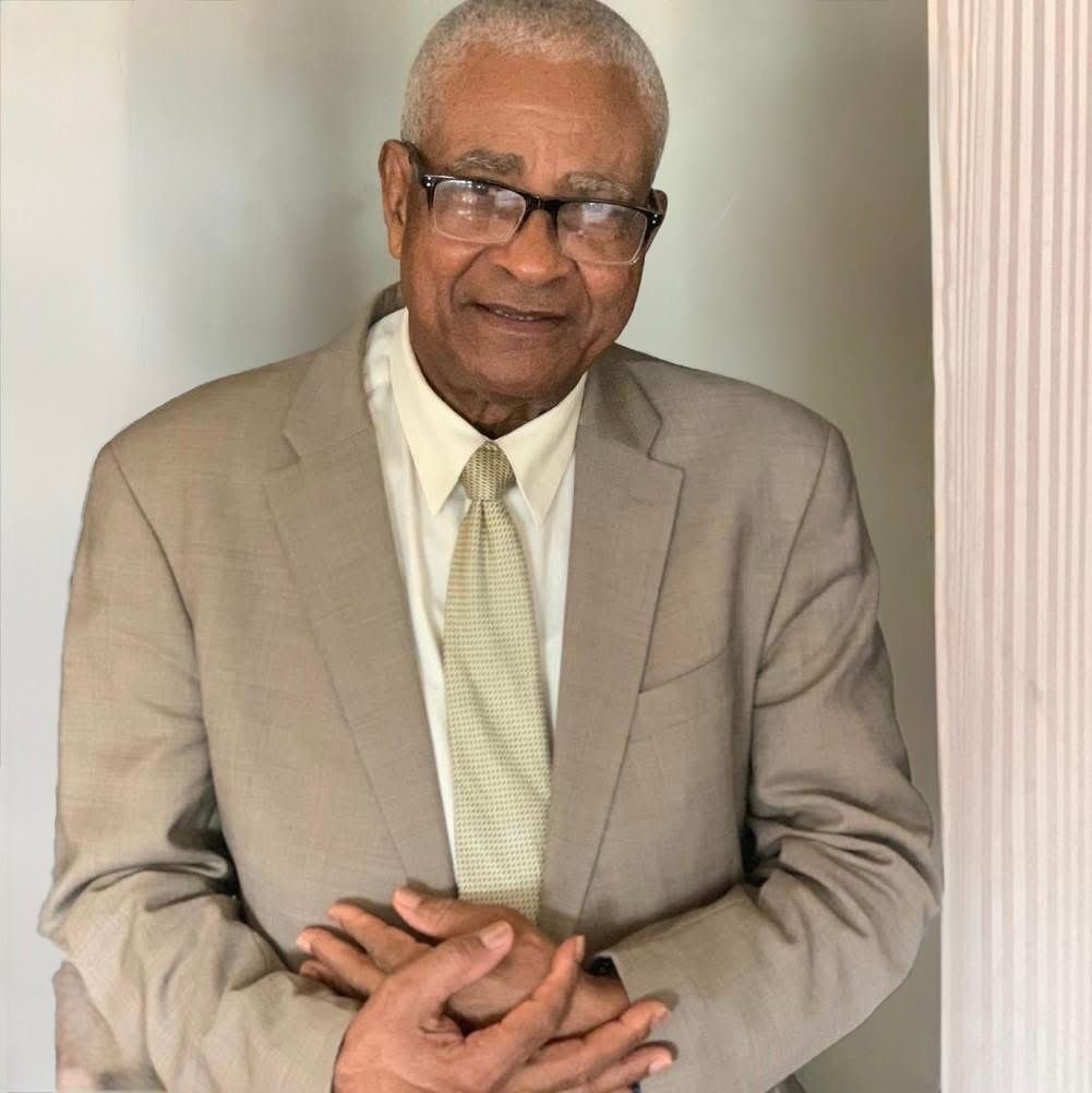 Older Black man in a tan suit and glasses, smiling, with hands clasped.