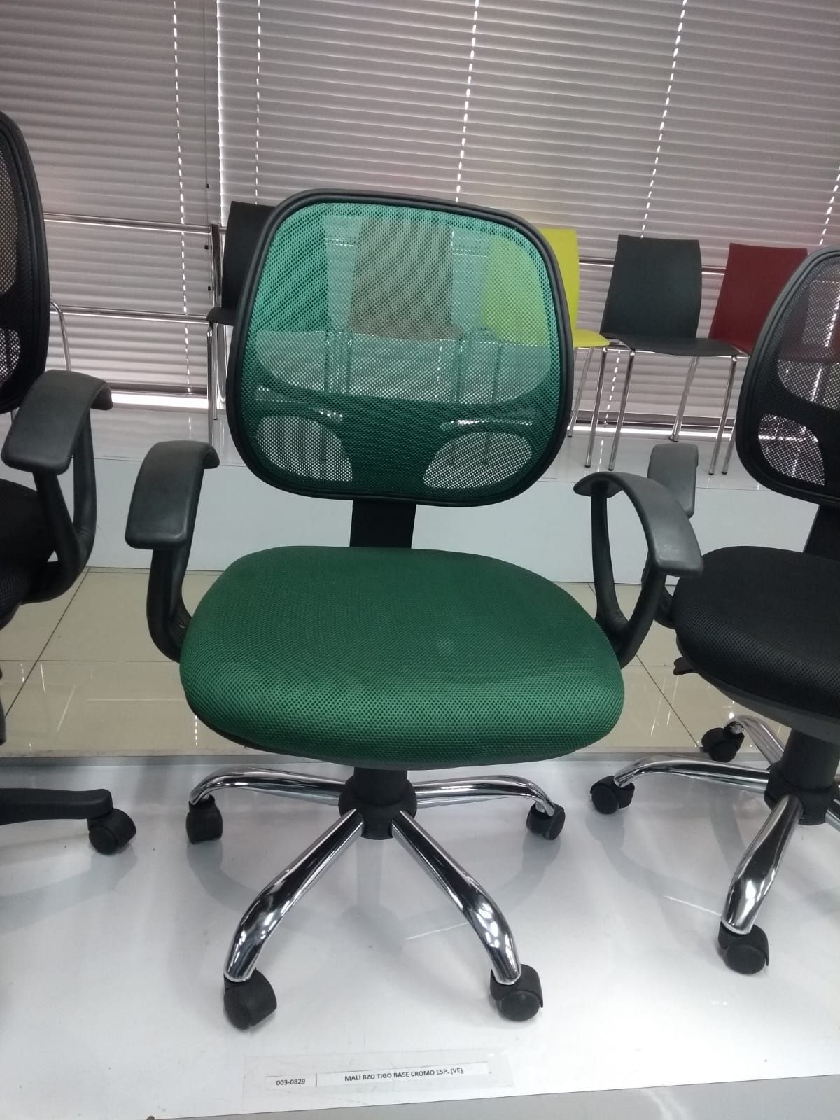 Silla de oficina de malla verde con base con ruedas en una sala de exposición, flanqueada por sillas negras.