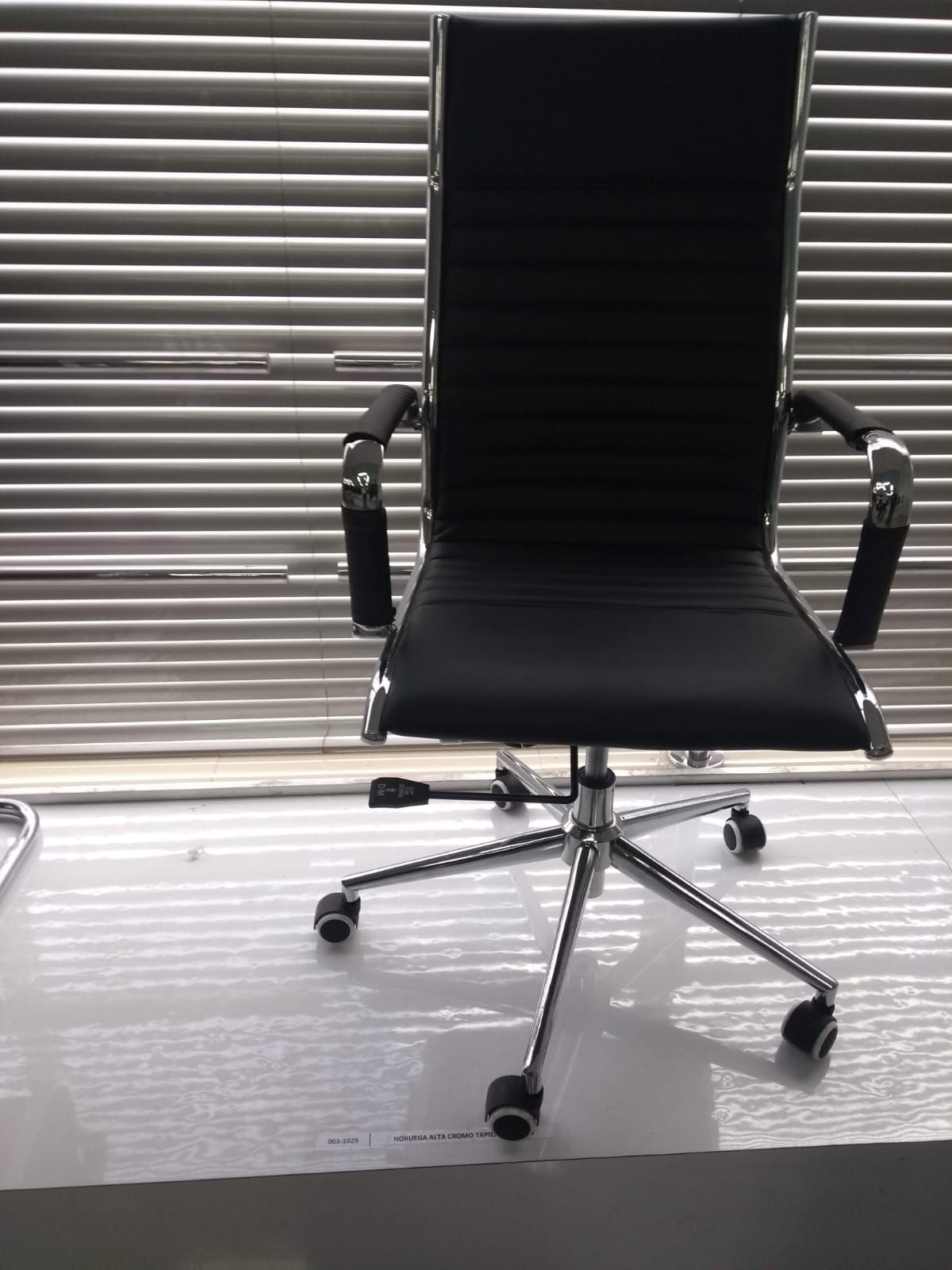 Silla de oficina negra con base cromada frente a persianas cerradas, sobre un suelo gris.