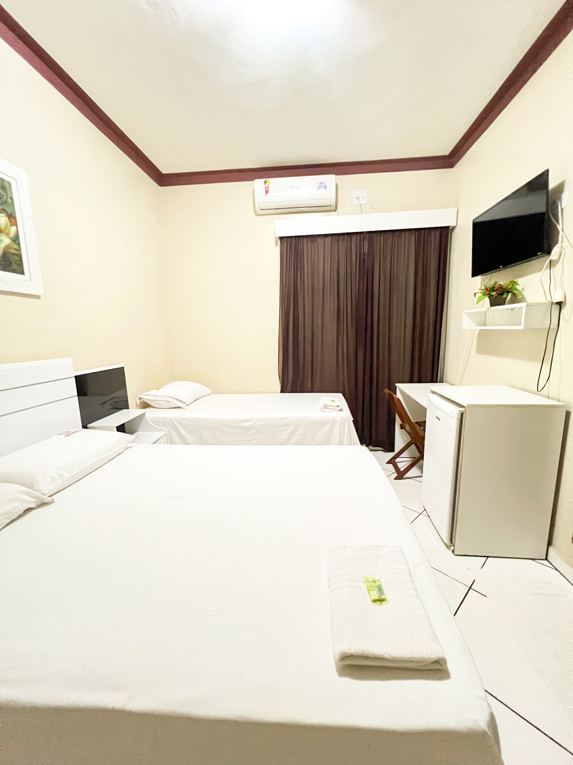 Quarto de hotel pequeno com duas camas brancas, TV de parede, ar condicionado e cortinas fechadas.