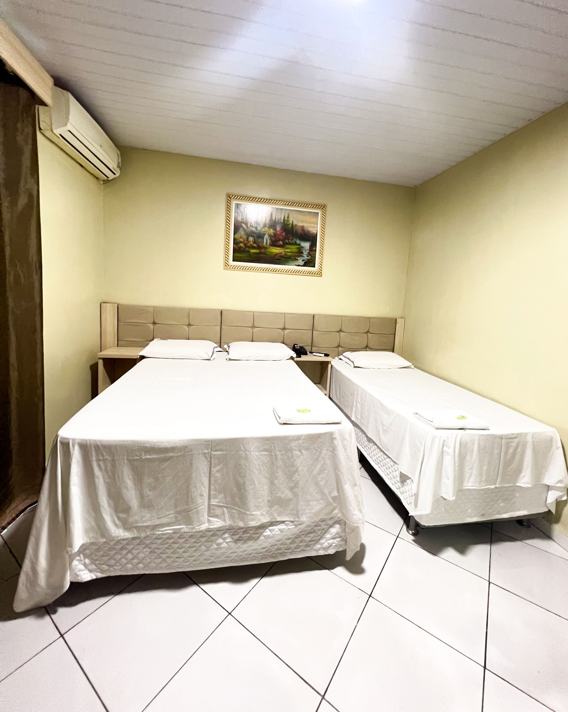 Quarto pequeno com duas camas de solteiro, lençóis brancos, piso de azulejo, obras de arte na parede e ar condicionado.