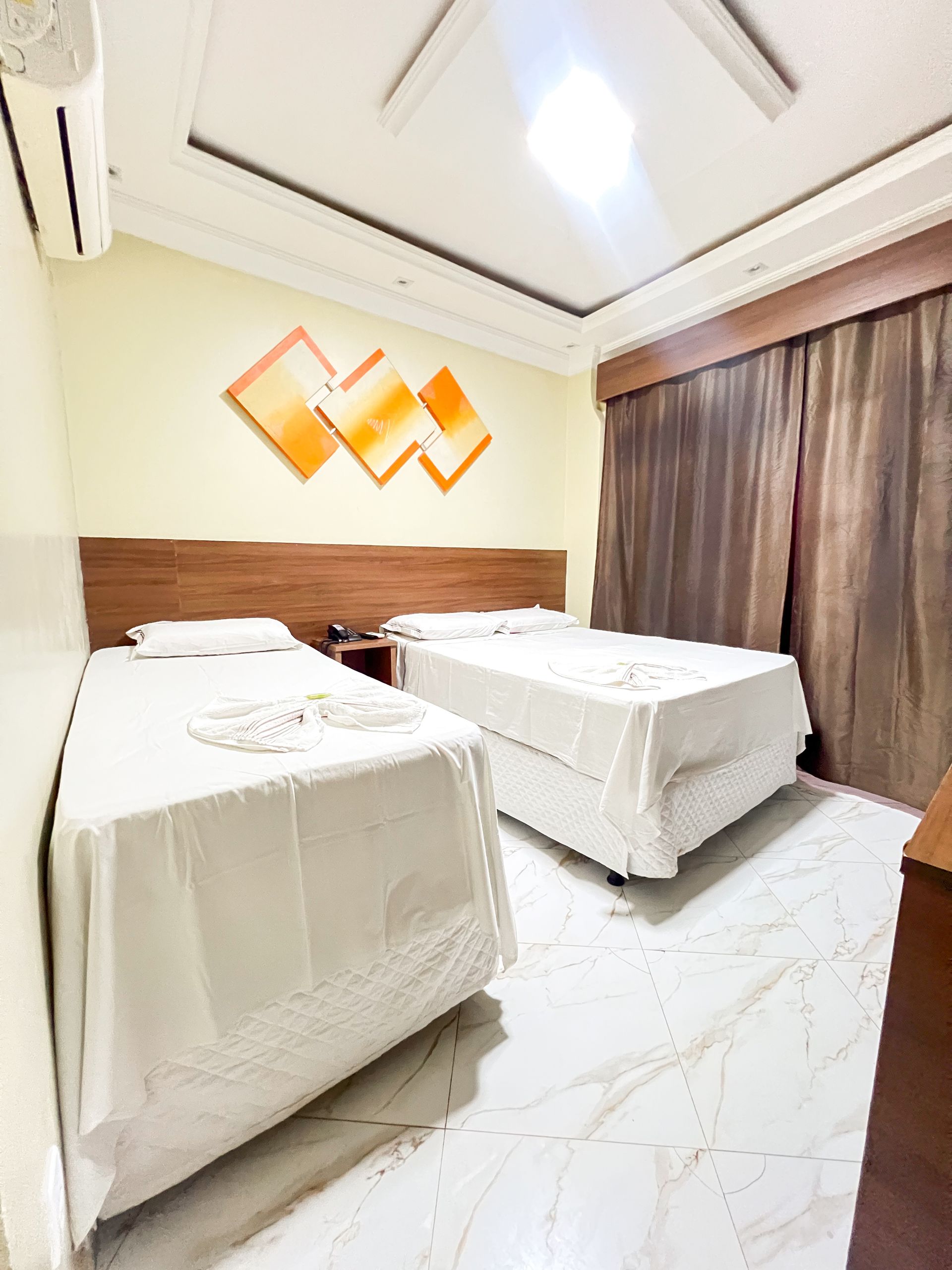 Quarto de hotel luminoso com duas camas de solteiro, roupa de cama branca, cabeceira de madeira e cortinas marrons.