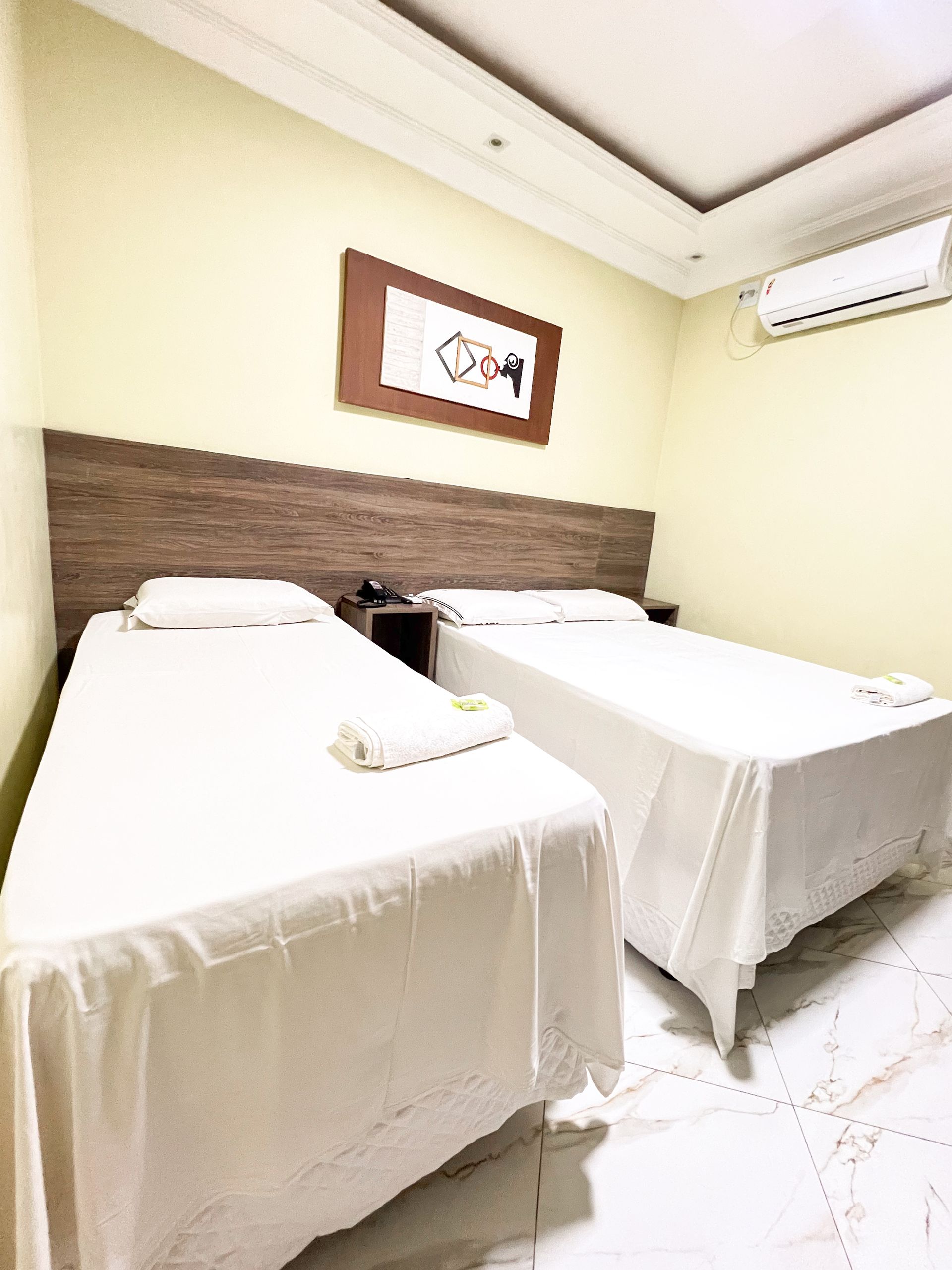 Quarto de hotel pequeno com duas camas de solteiro bem arrumadas, cabeceira de madeira e obras de arte na parede.