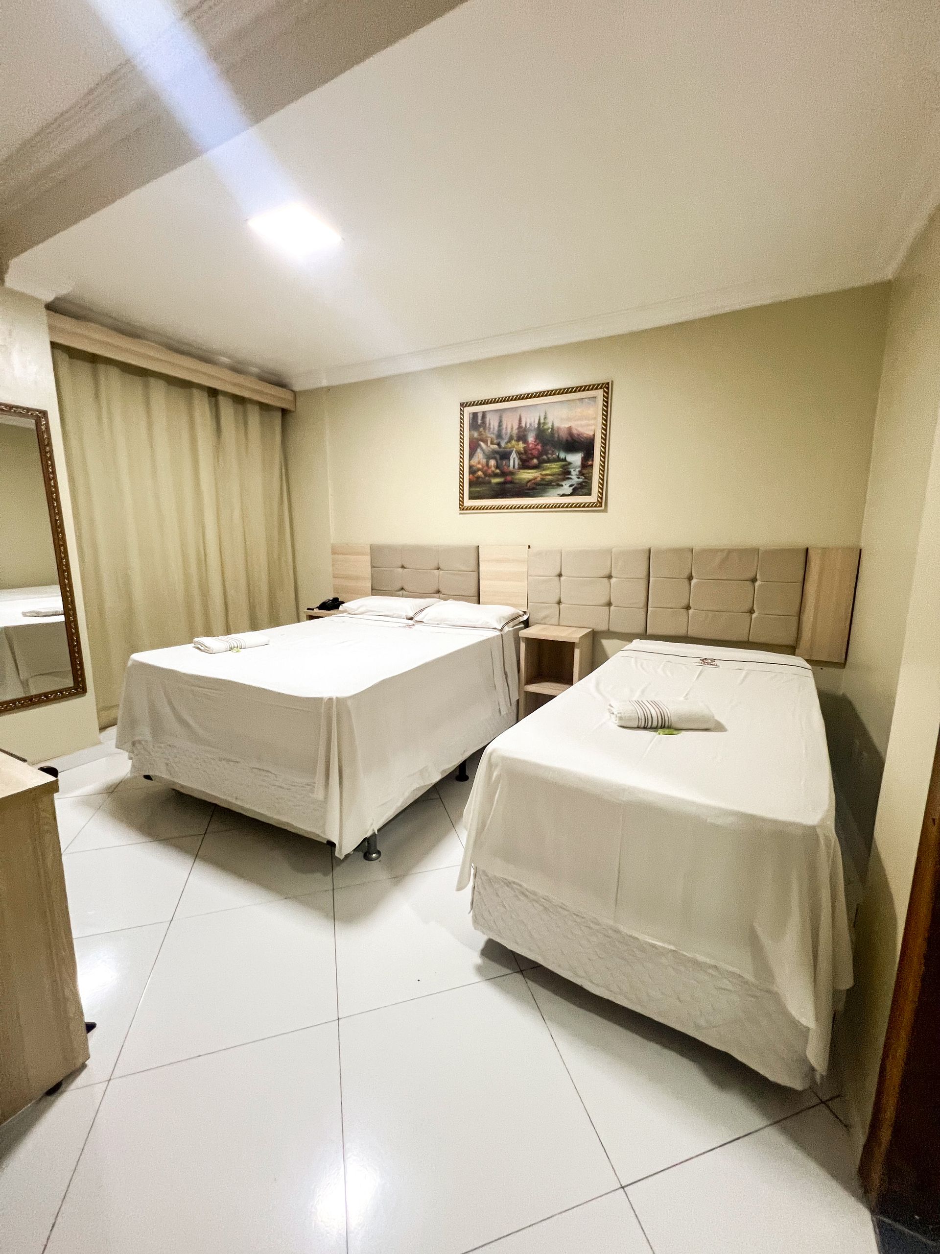 Quarto de hotel impecável com duas camas de solteiro, paredes bege, piso de azulejo branco e um quadro emoldurado acima das cabeceiras.