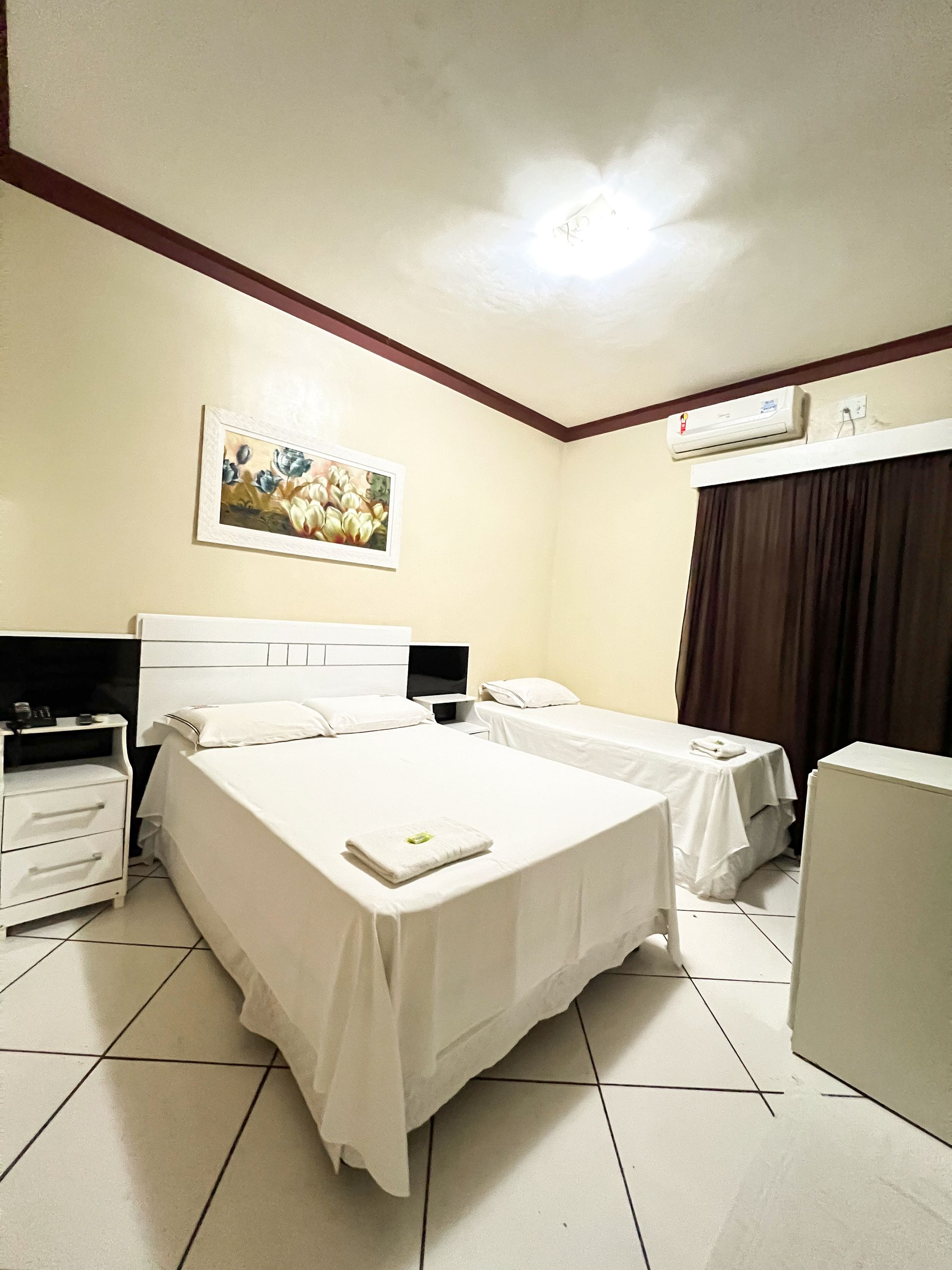 Quarto de hotel pequeno com duas camas de solteiro, lençóis brancos, cortinas escuras e piso de azulejo.