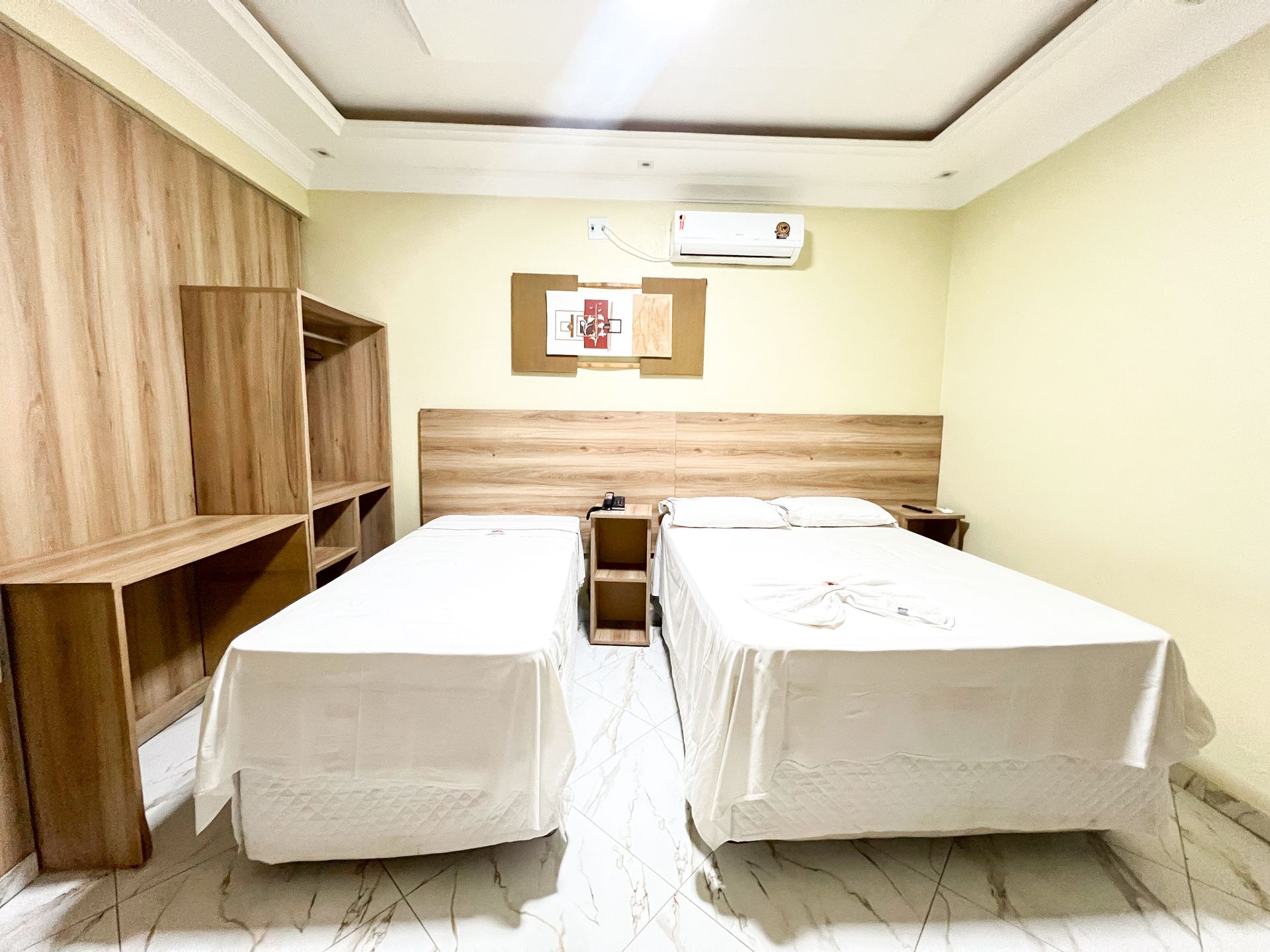Quarto de hotel pequeno com duas camas de solteiro, painéis de madeira, uma escrivaninha e um ar-condicionado de parede.