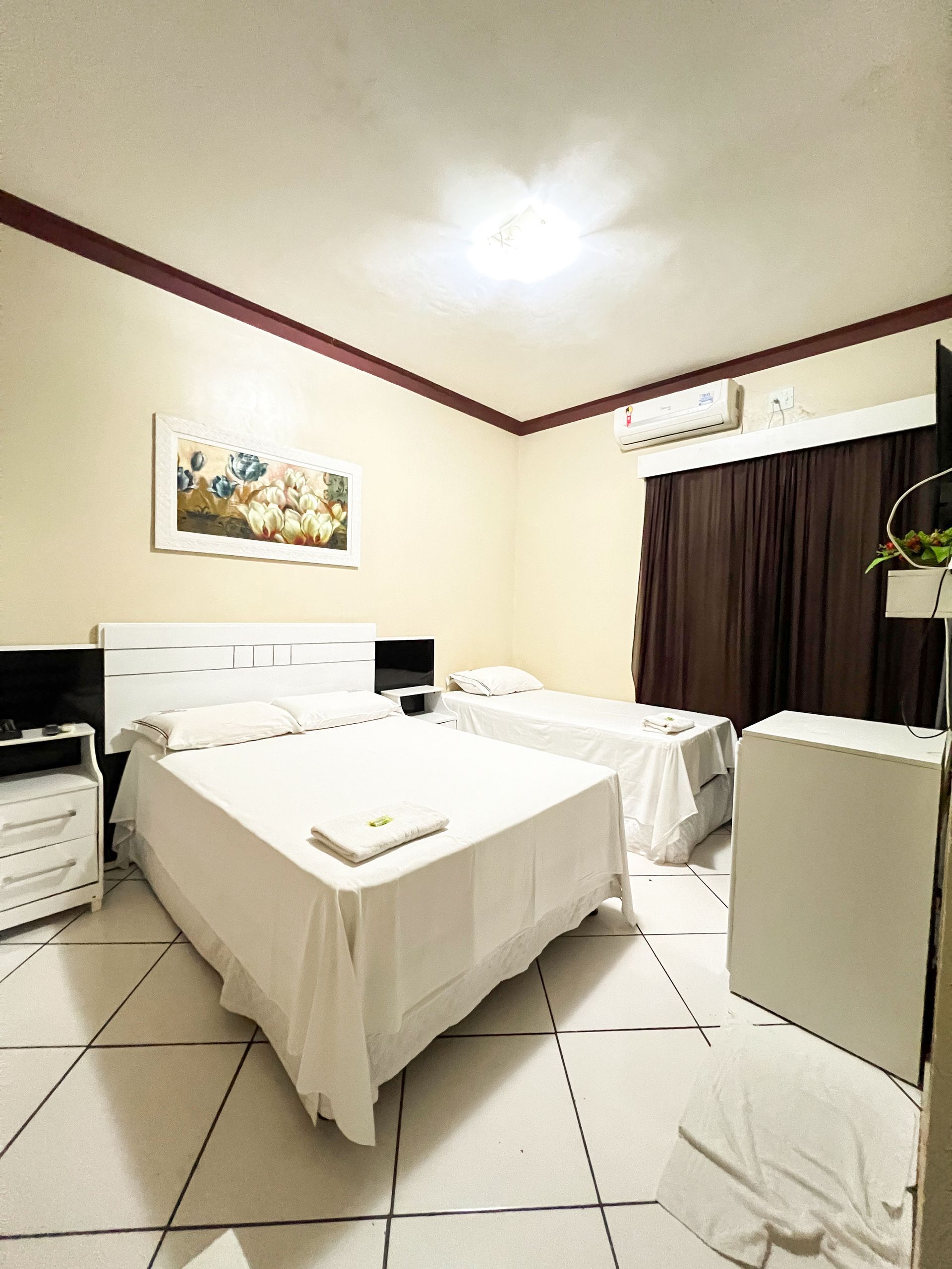 Quarto de hotel pequeno e branco com duas camas de solteiro, piso de azulejo, cortinas escuras e ar-condicionado de parede.