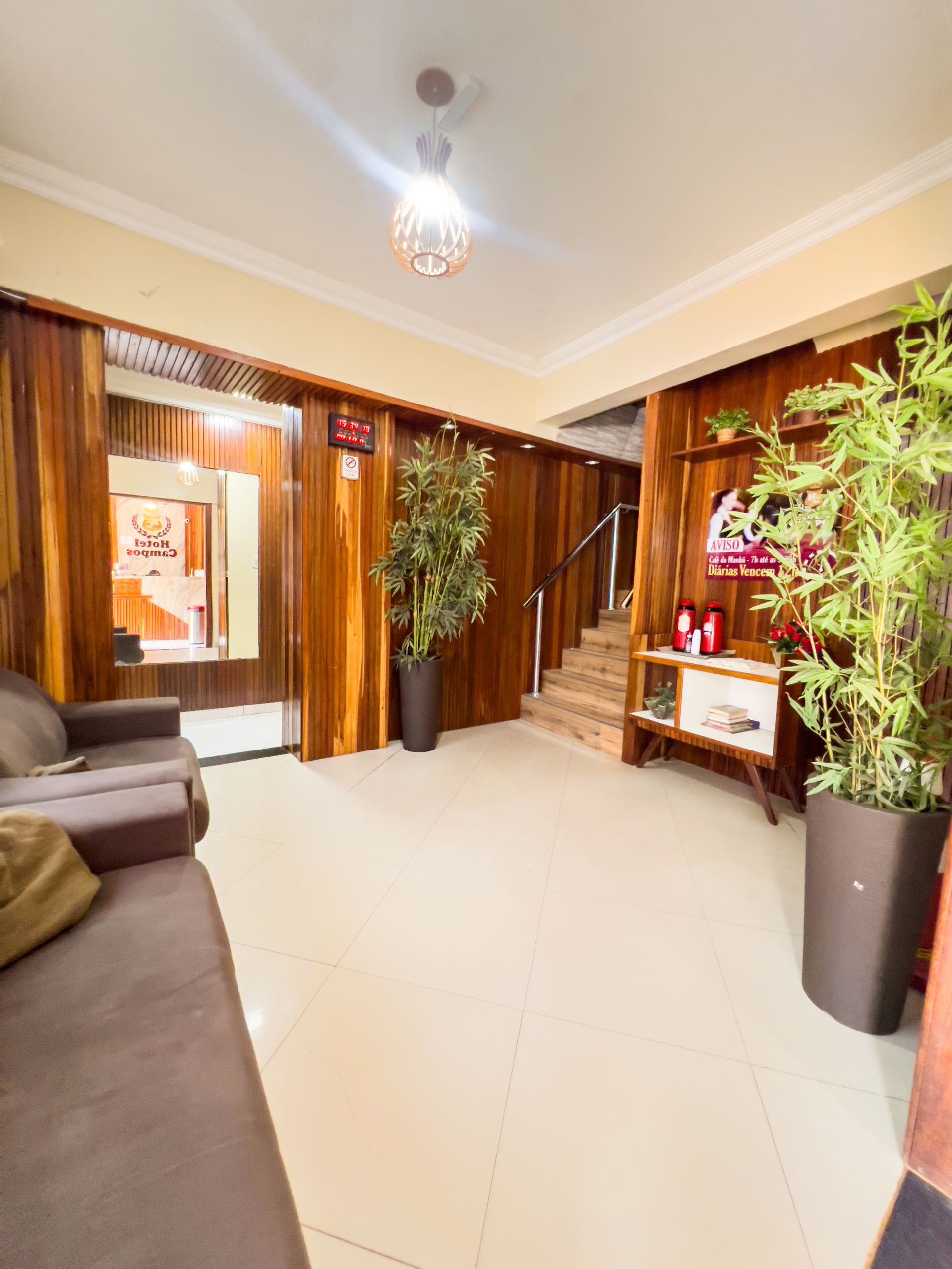 Sala de estar iluminada com sofá marrom, plantas altas e portas de madeira sobre piso de azulejo.
