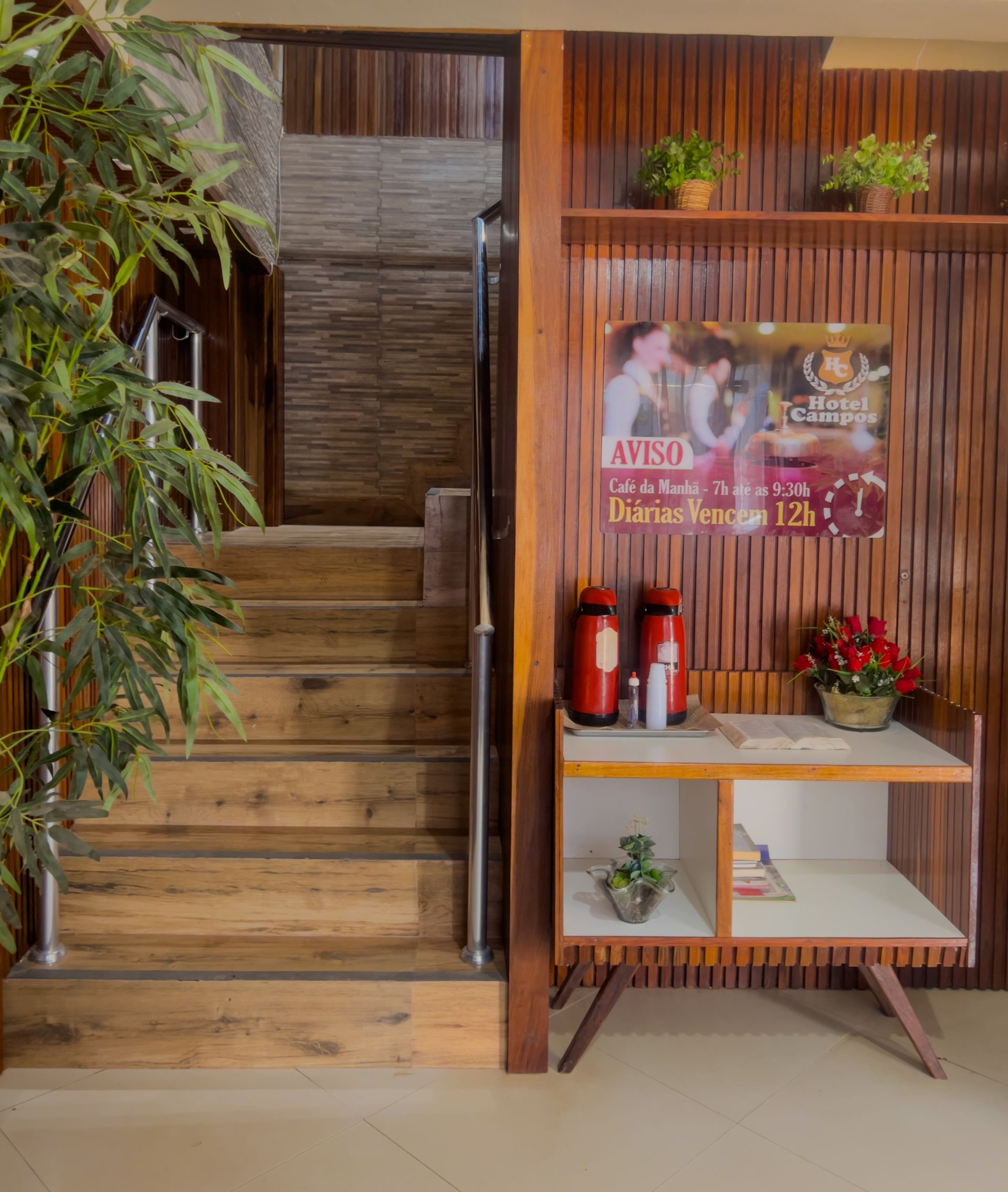 Hall de entrada de madeira com escadas, vasos de plantas e uma pequena mesa de exposição com pôsteres e flores.