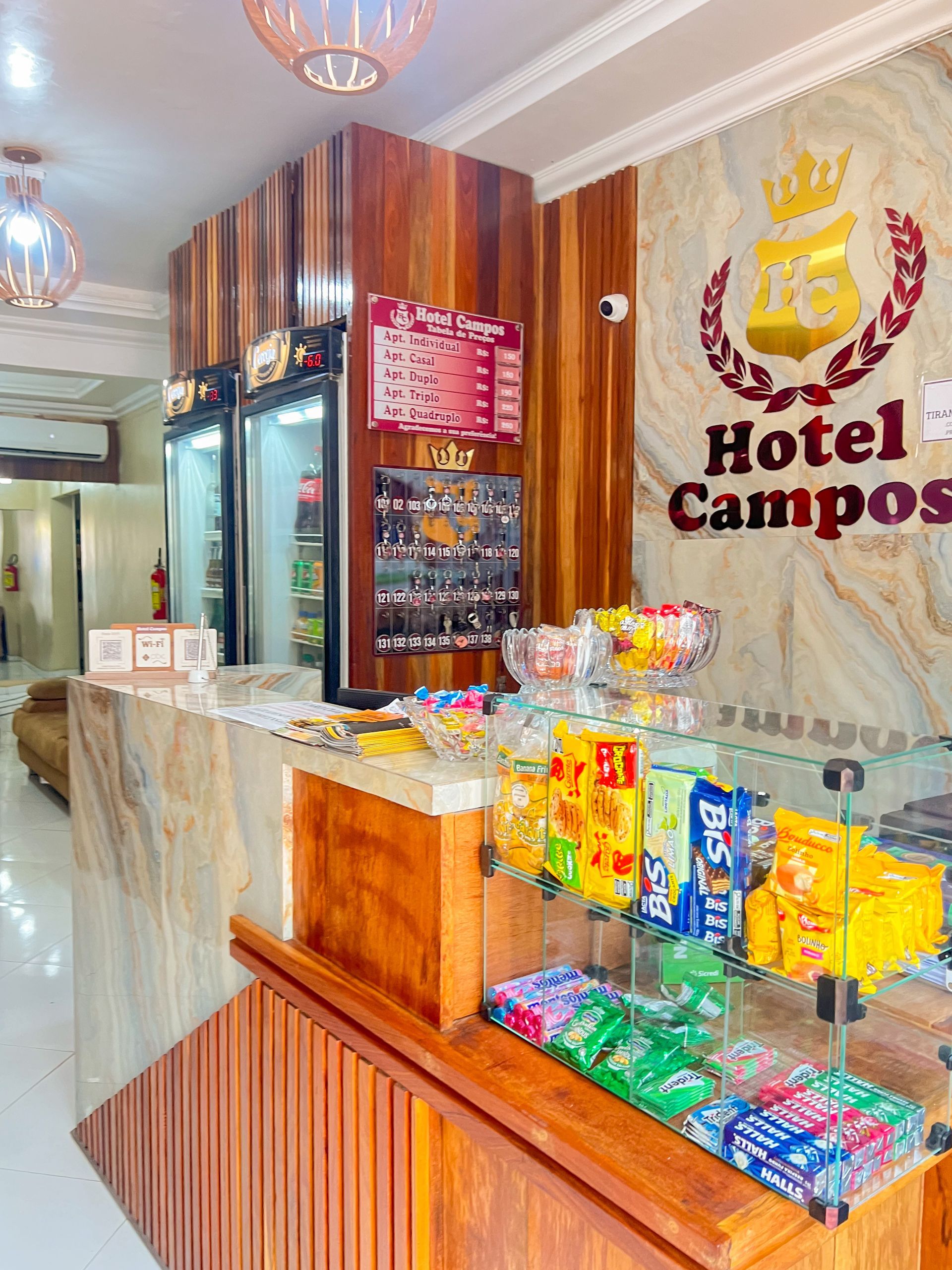 Balcão de recepção do Hotel Campos com balcão de mármore, expositor de lanches e logotipo na parede em um lobby iluminado.