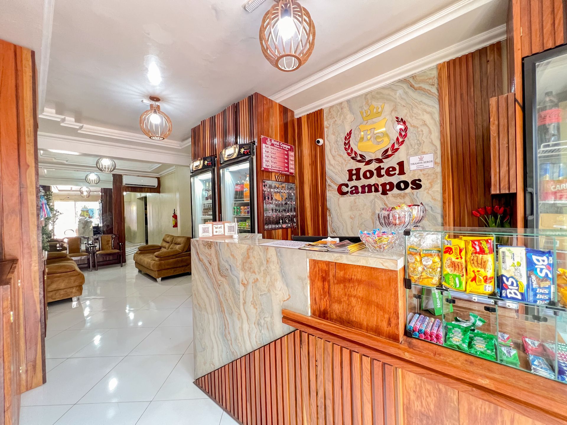 Balcão de recepção do hotel com fachada de mármore, painéis de madeira, expositor de lanches e área de estar em um saguão iluminado.