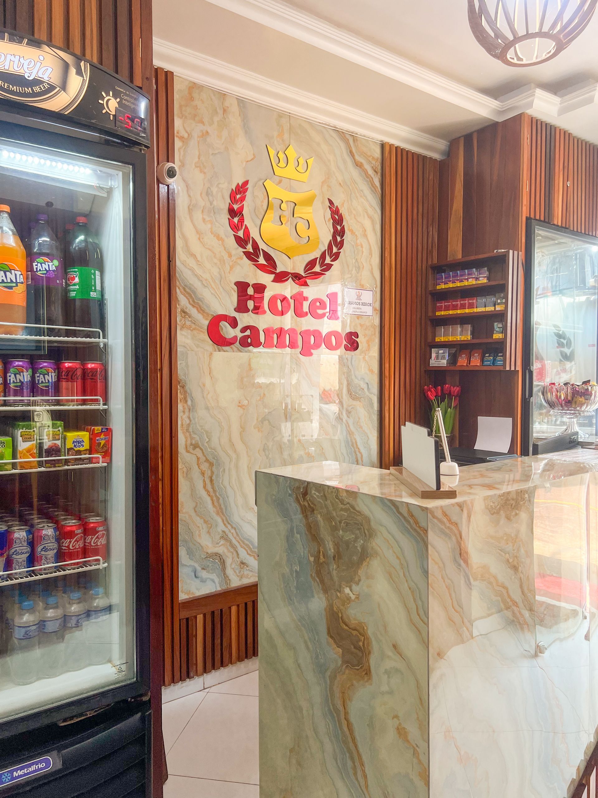 Lobby do Hotel Campos com balcão de recepção em mármore, painéis de madeira e refrigeradores de bebidas na entrada.
