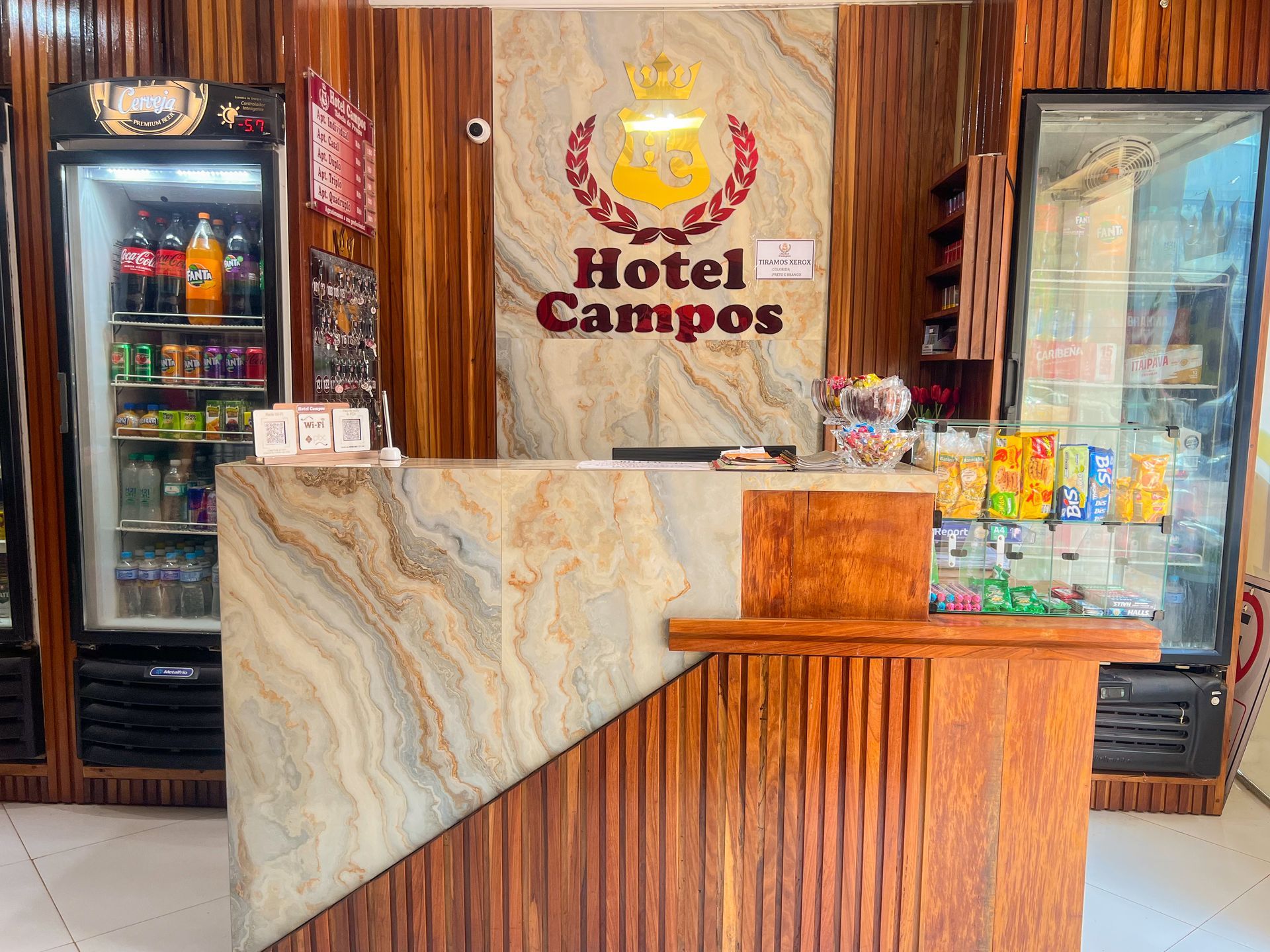 Balcão de recepção do Hotel Campos com painéis de madeira, balcão de mármore, refrigeradores de bebidas e placa com o logotipo atrás.