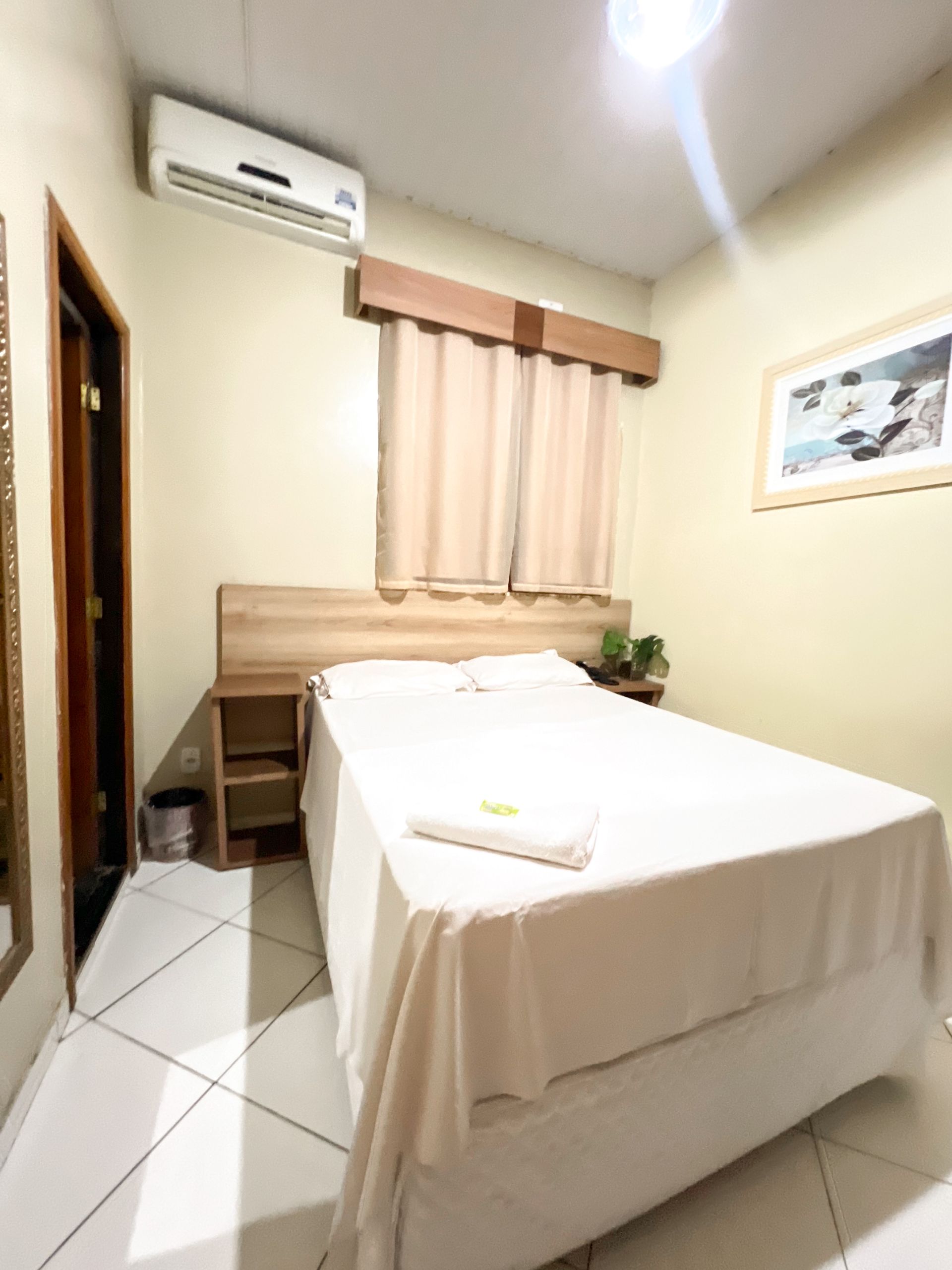 Quarto pequeno com cama, cortinas na janela, ar condicionado de parede e piso de azulejo.