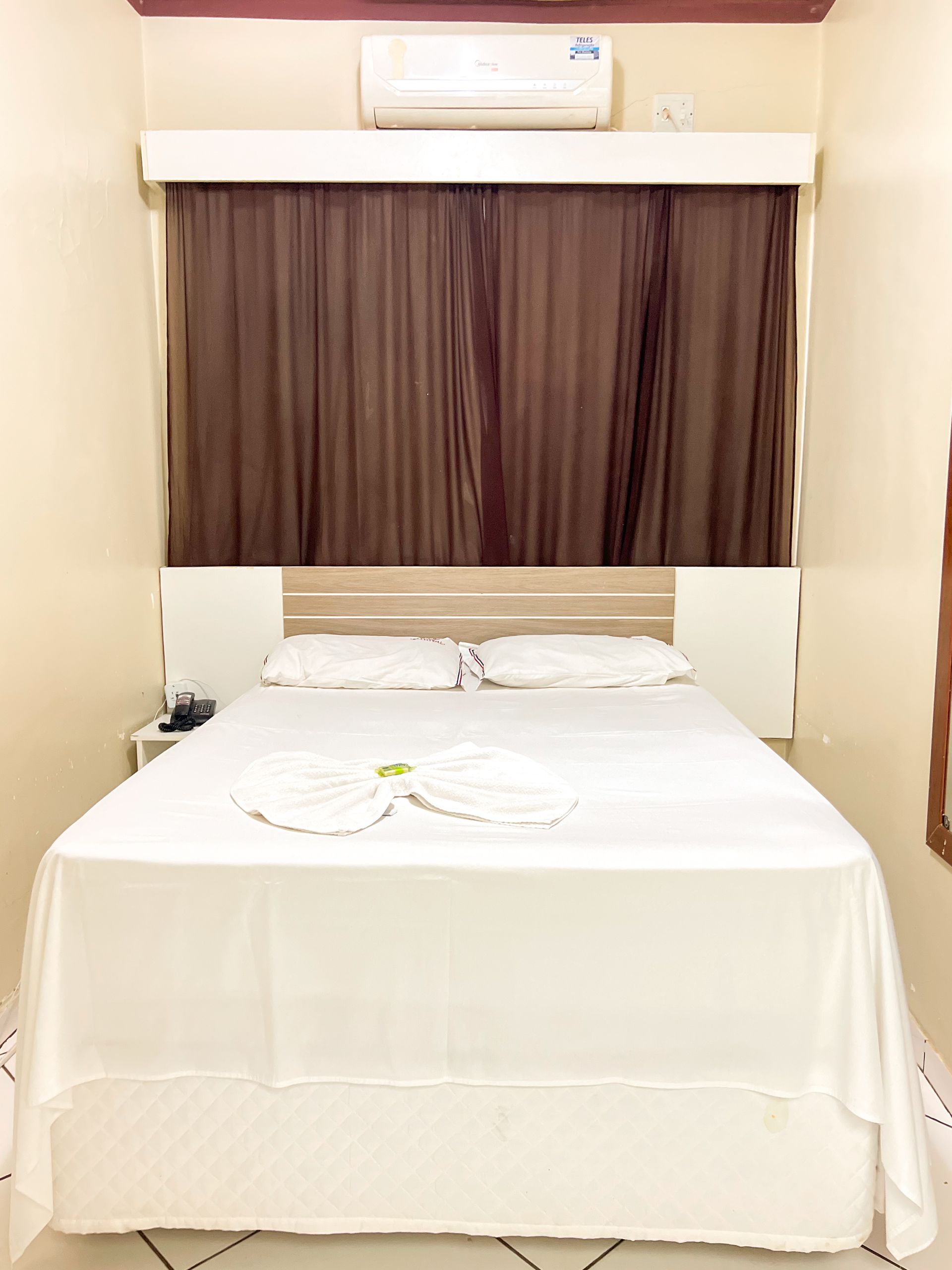 Uma cama bem arrumada em um pequeno quarto de hotel com cortinas marrons e um ar-condicionado acima dela.