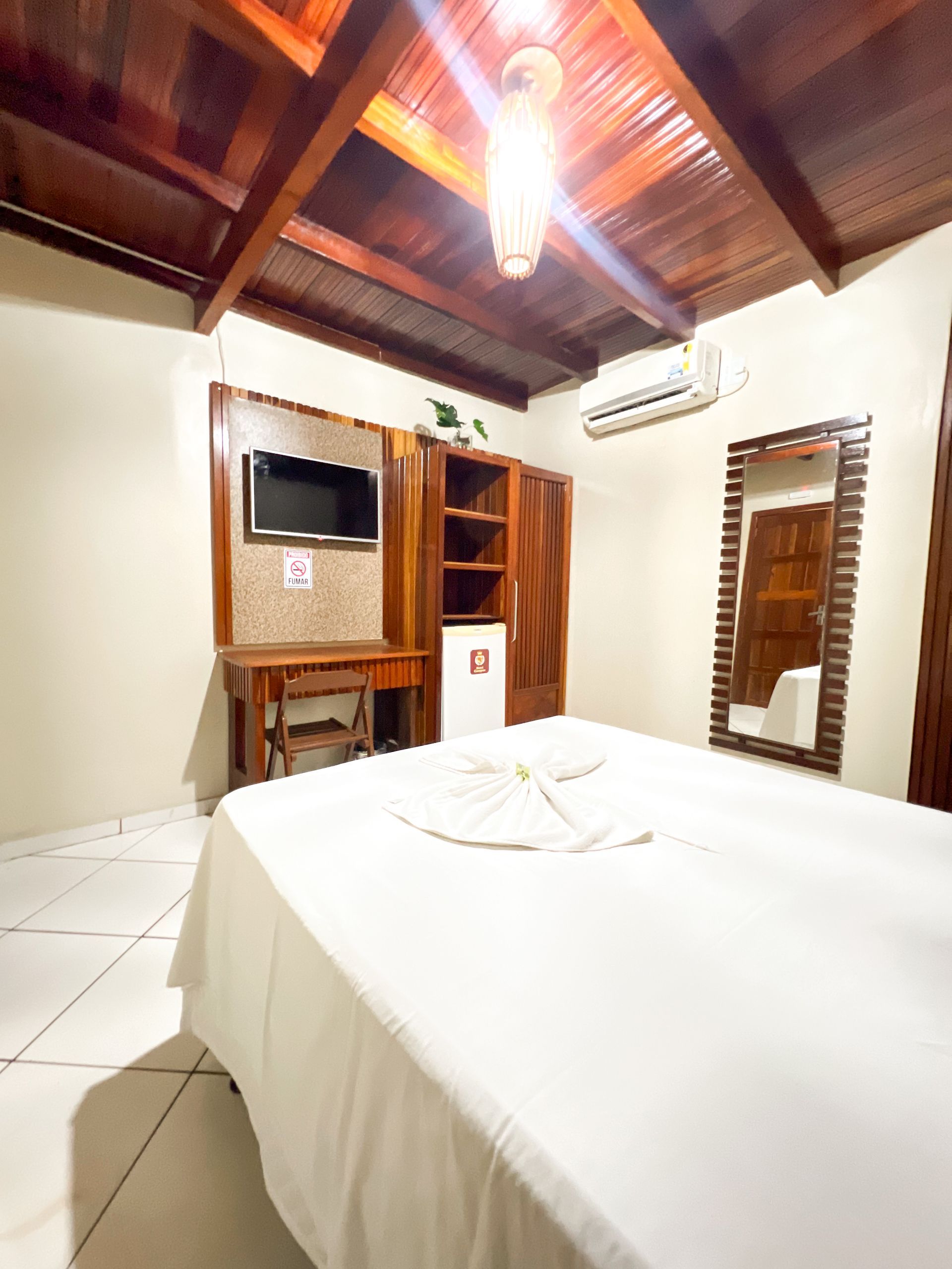 Quarto de hotel luminoso com cama branca, teto de madeira, TV, guarda-roupa e espelho de parede.