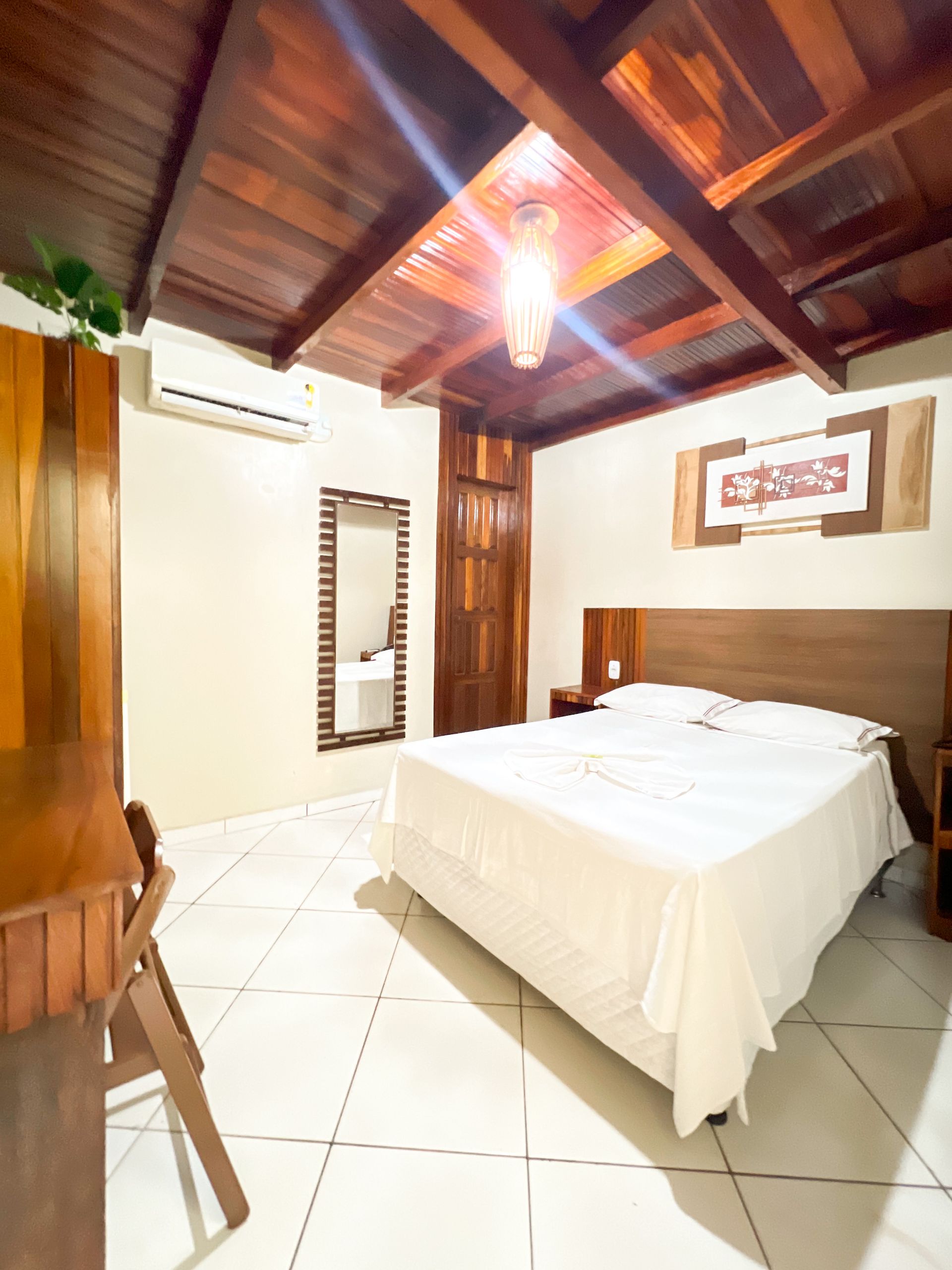 Quarto rústico com teto de madeira, cama branca, piso de azulejo e obras de arte na parede.