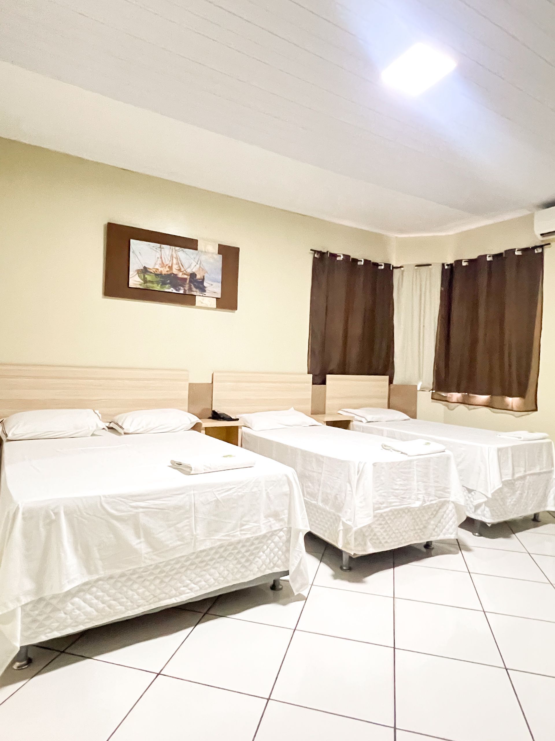 Quarto de hotel com três camas de solteiro, lençóis brancos, cortinas marrons e piso de azulejo.