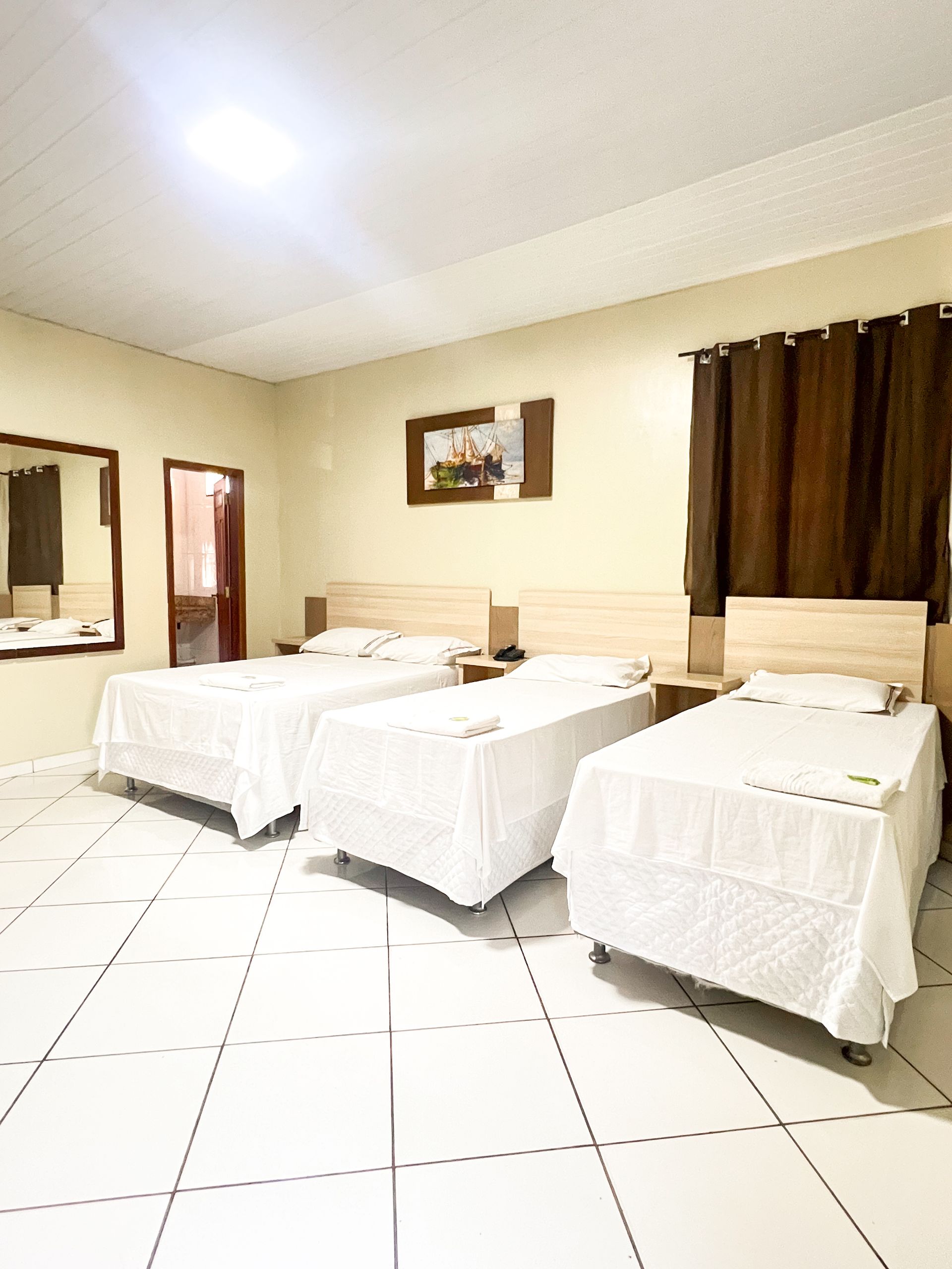 Quarto de hotel simples com três camas de solteiro, roupa de cama branca, um espelho e cortinas marrons.